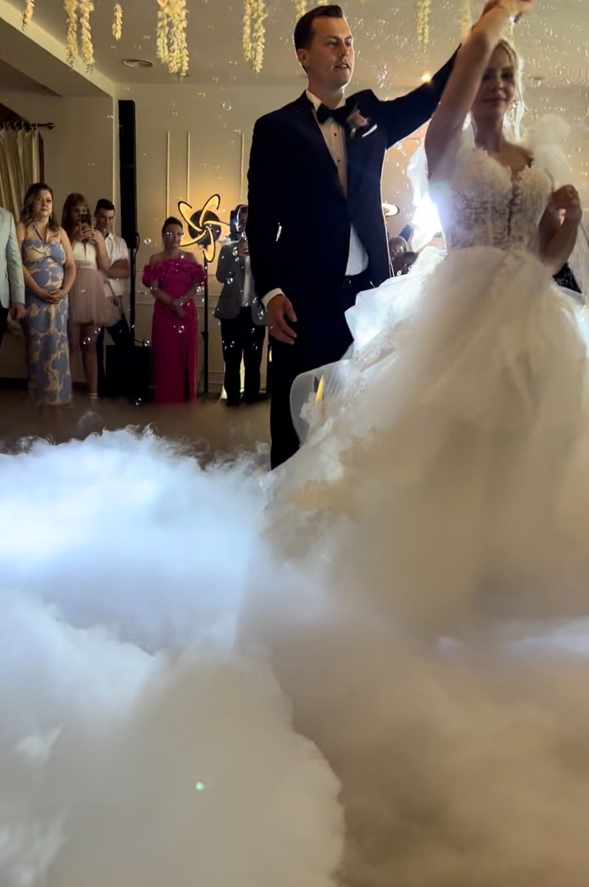 Para młoda w tańcu, otoczona efektem ciężkiego dymu na parkiecie. Goście weselni w tle, dekoracje kwiatowe i oświetlenie tworzą romantyczny nastrój.