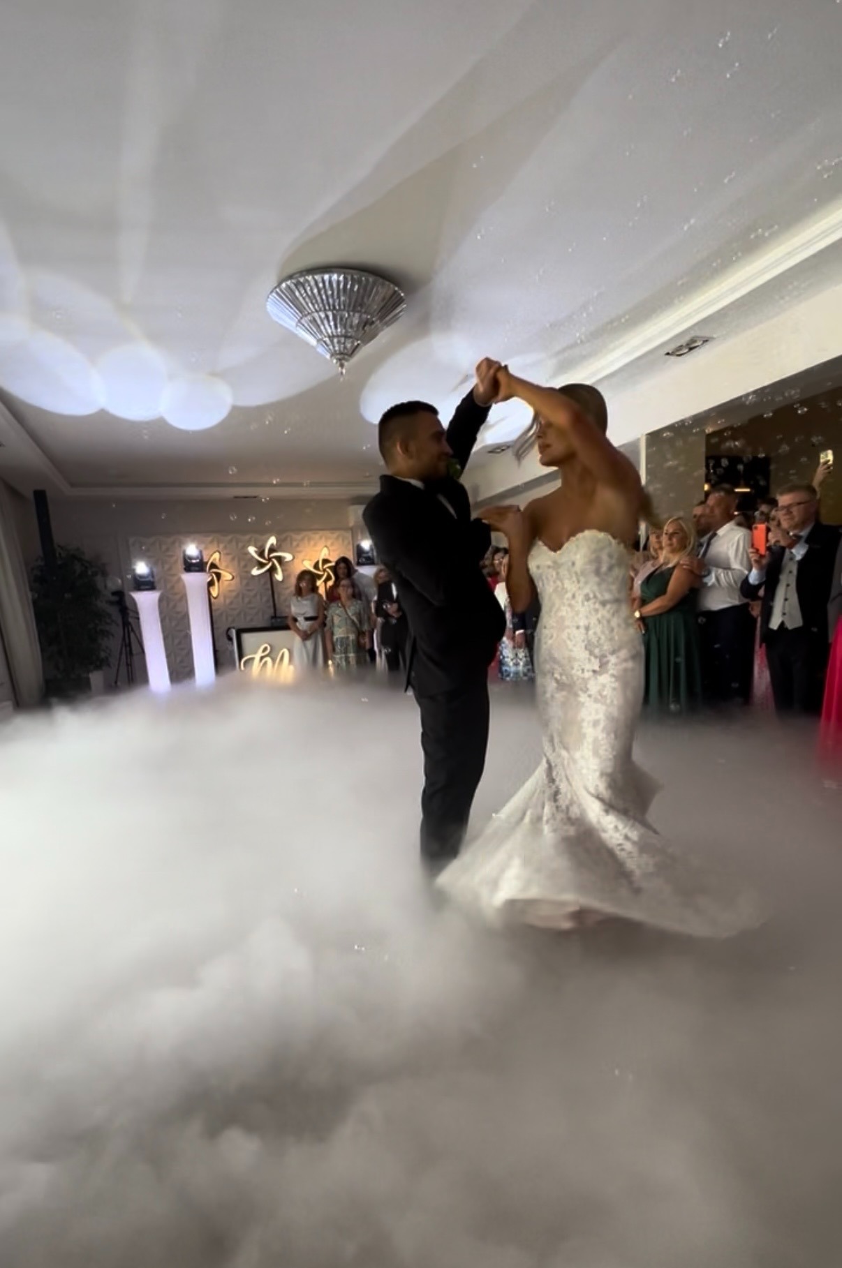 Para młoda tańczy pierwszy taniec w dymie na sali weselnej z dekoracjami świetlnymi i gośćmi w tle. Romantyczna atmosfera.