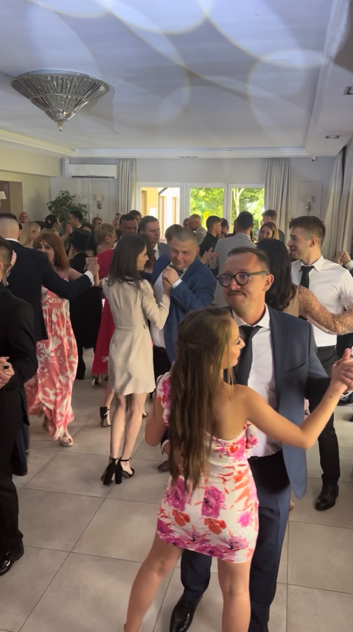 Parkiet taneczny wypełniony gośćmi weselnymi w eleganckich strojach, tańczącymi w jasnym, przestronnym wnętrzu sali balowej z żyrandolem.