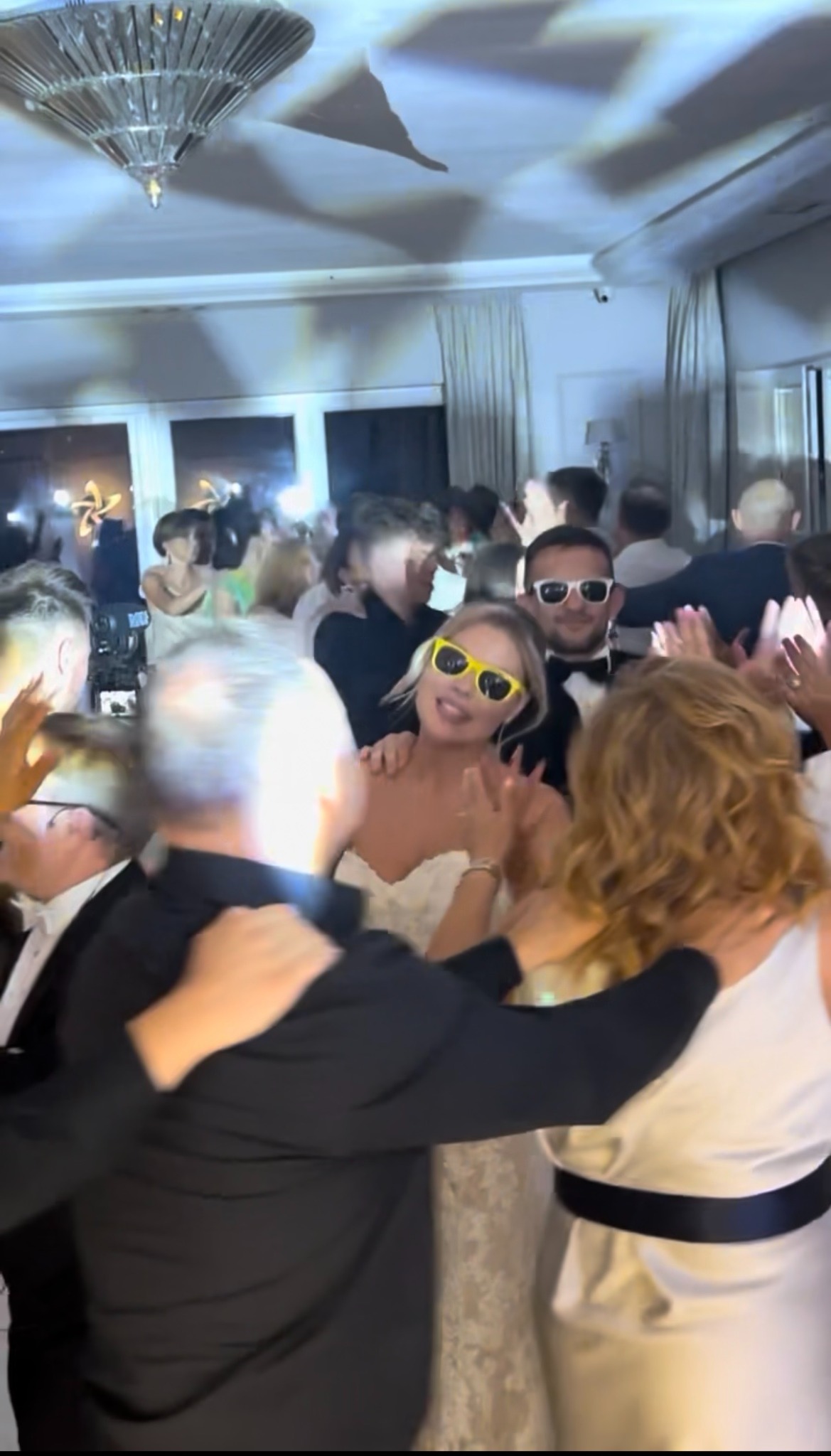 Roztańczona para młoda w okularach na parkiecie, otoczona gośćmi weselnymi w eleganckim wnętrzu z kryształowym żyrandolem. Radosna atmosfera.