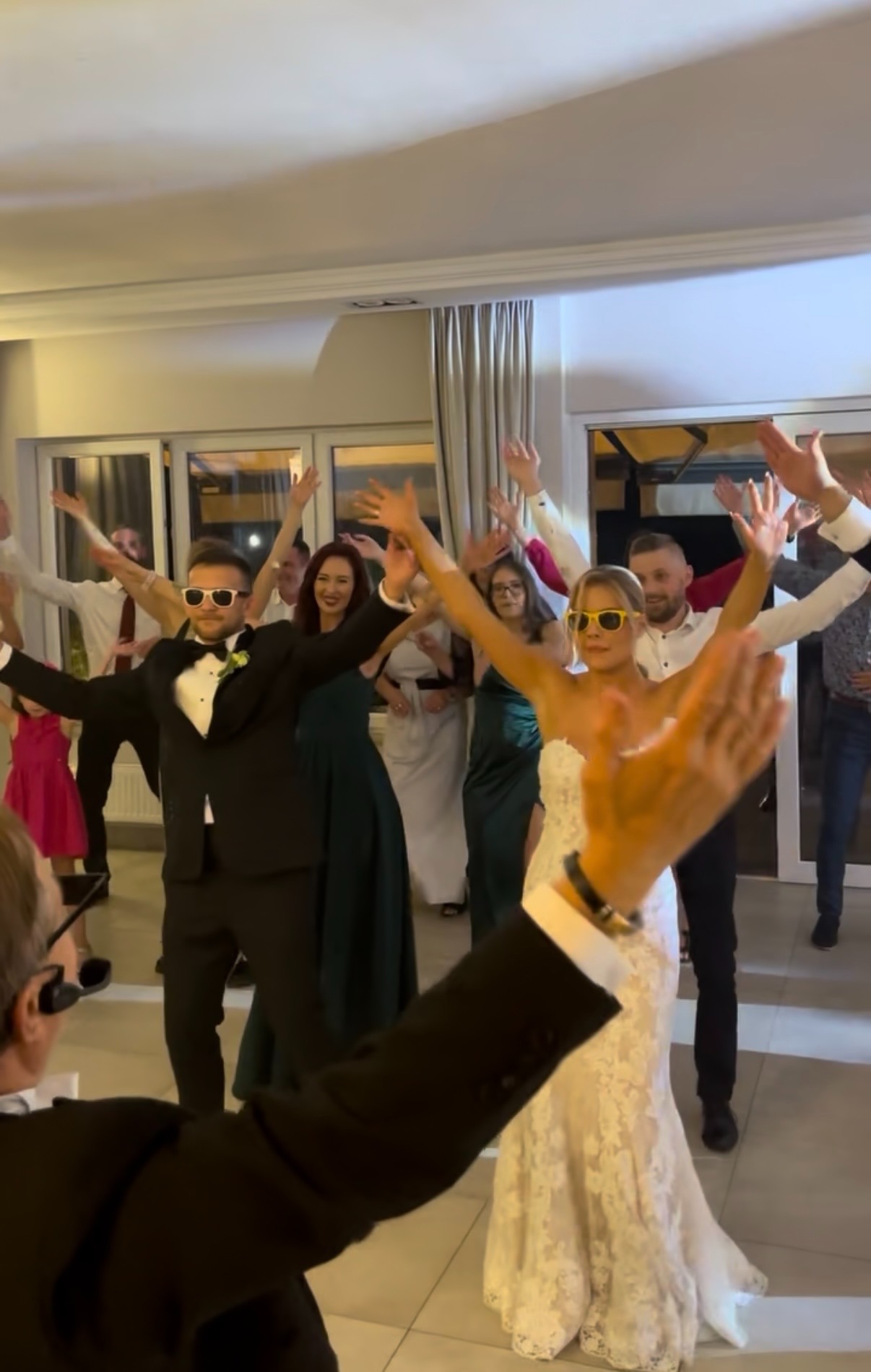 Rozbawieni goście weselni w okularach tańczą z podniesionymi rękami, w tle para młoda. Zabawa weselna w sali z oknami.