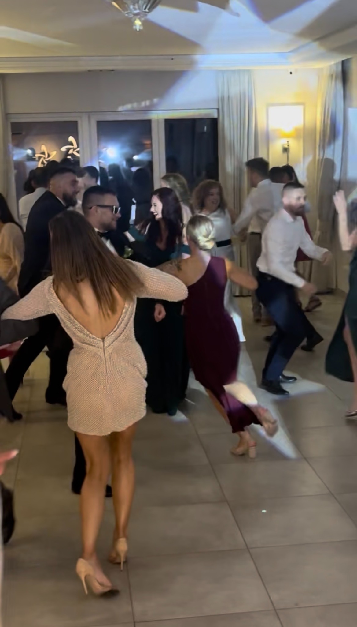 Roztańczona grupa gości weselnych w eleganckiej sali balowej, w tle okna z białymi zasłonami i dekoracyjne oświetlenie.