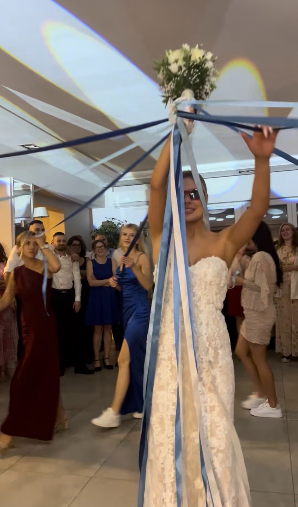 Panna młoda w koronkowej sukni, z bukietem i wstążkami, w trakcie weselnego oczepiny, w tle goście i dekoracje sali.