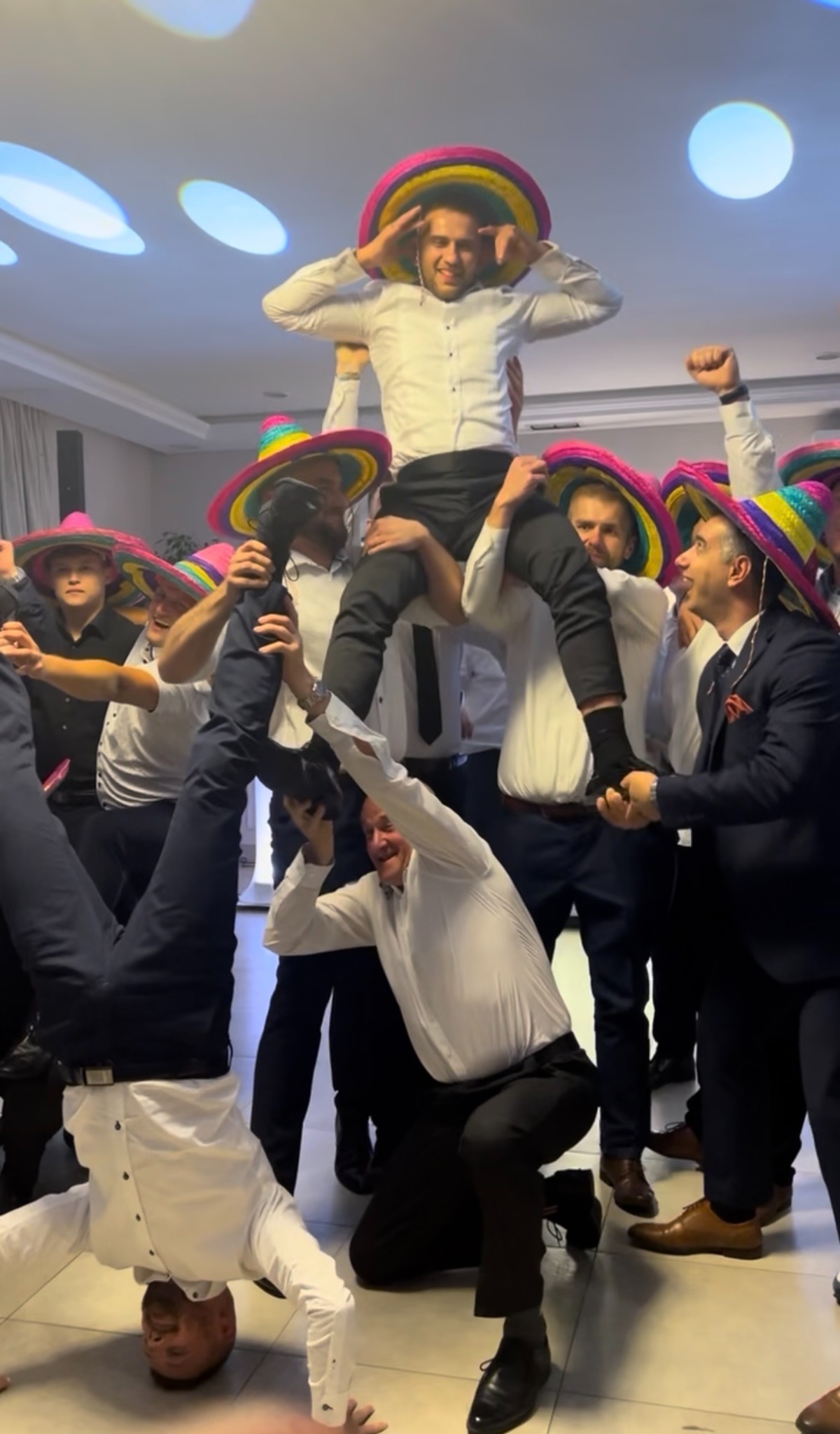Rozbawiona grupa mężczyzn w kolorowych sombrerach świętuje, jeden na barana, inny robi stójkę na głowie. Radosna atmosfera imprezy.
