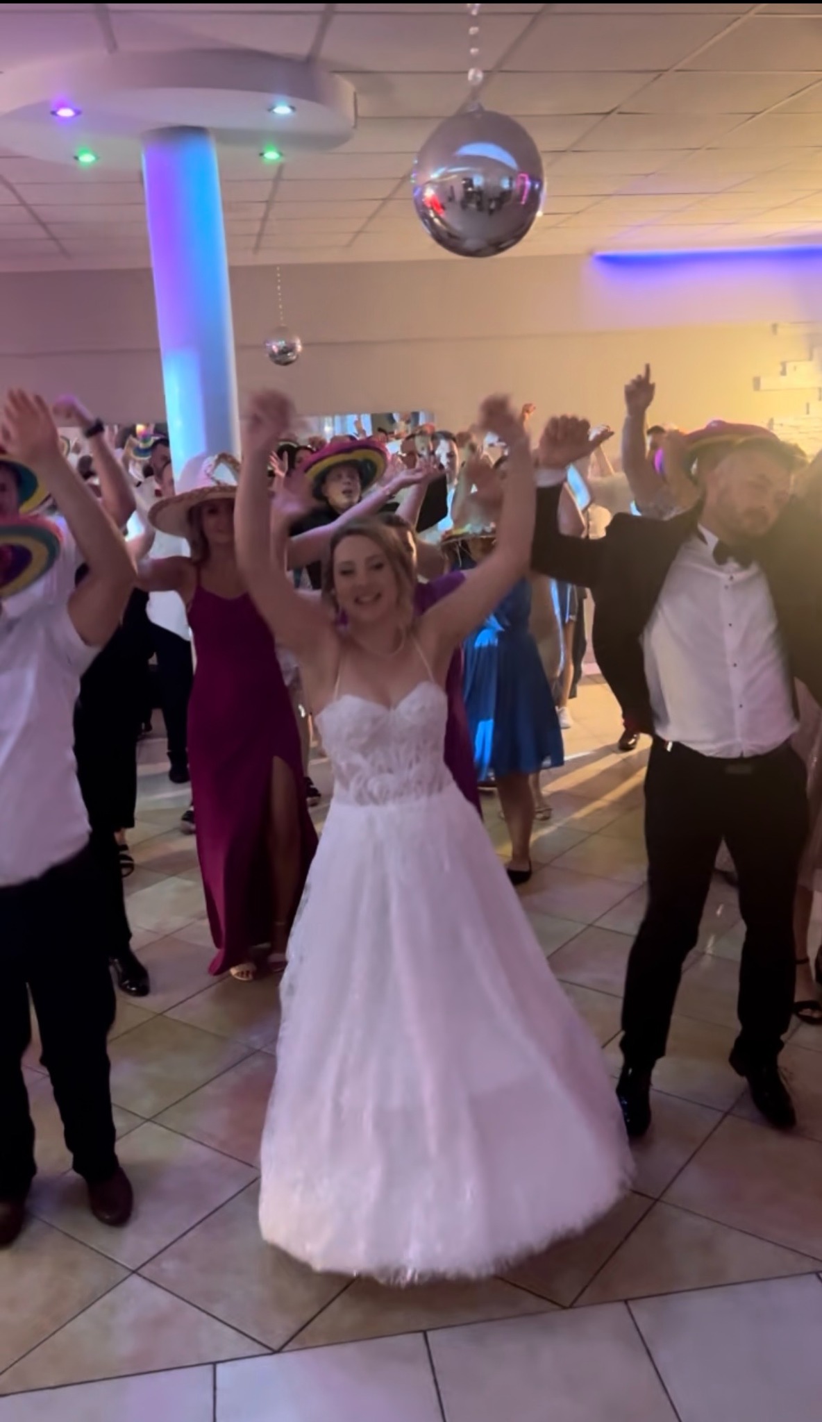 Rozbawieni goście weselni w tańcu pod srebrną kulą, w tym panna młoda w białej sukni i pan młody w garniturze. Kolorowe oświetlenie parkietu.