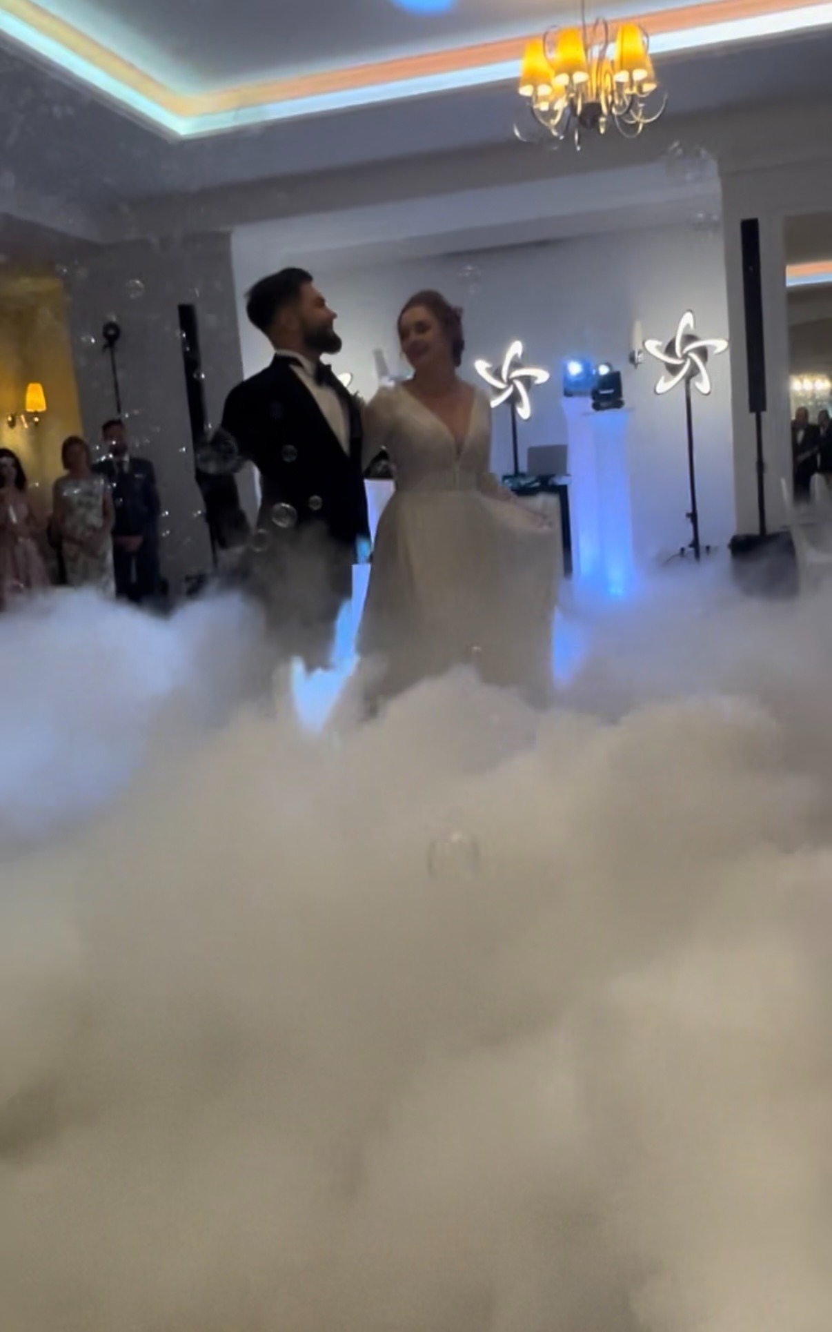 Para młoda tańczy w dymie na sali weselnej, w tle dekoracje świetlne i goście. Romantyczna atmosfera z efektem specjalnym.