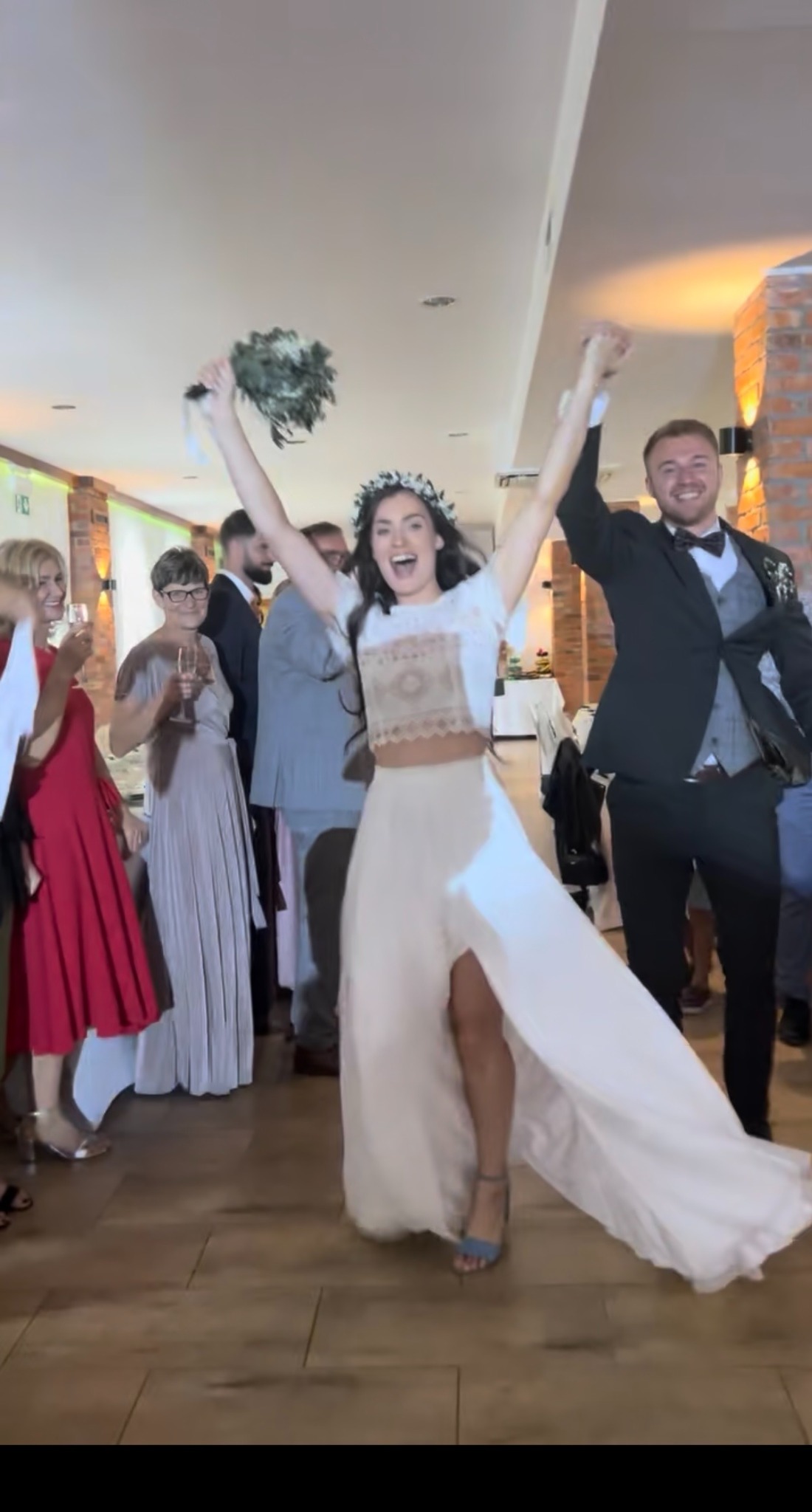 Radosna panna młoda z bukietem i pan młody świętują na sali weselnej, otoczeni gośćmi. Uśmiechy, uniesione ręce i taneczny nastrój.