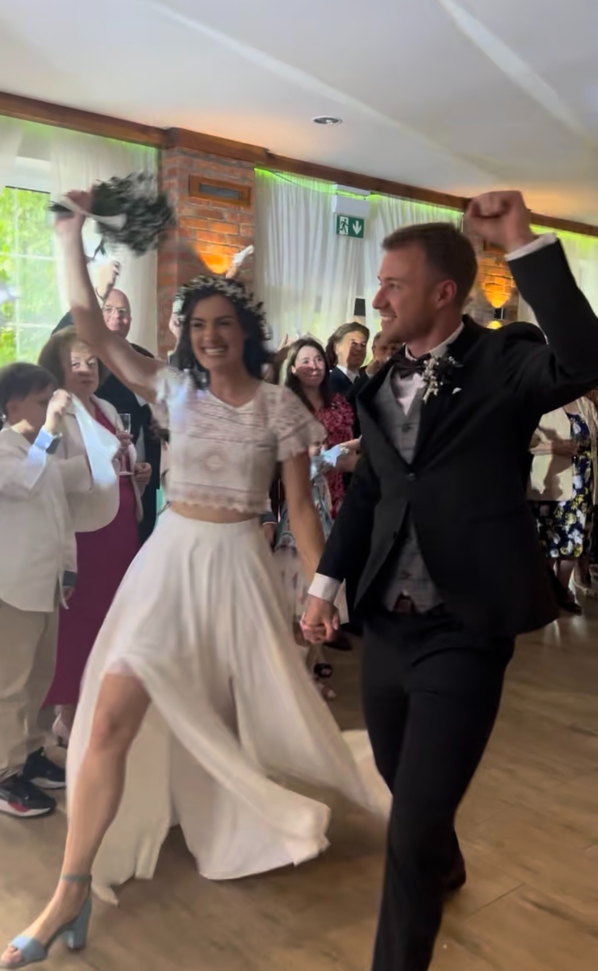 Uśmiechnięta para młoda, trzymając się za ręce, świętuje swój ślub w otoczeniu gości. Panna młoda w koronie i zwiewnej sukni, pan młody w eleganckim garniturze.