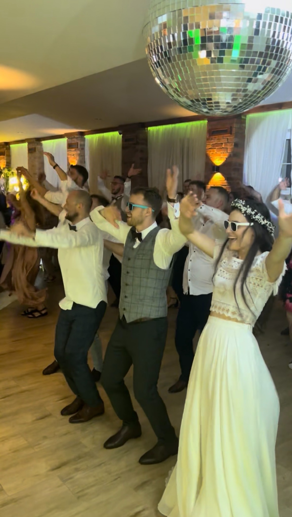 Roztańczona para młoda w białych okularach i goście weselni pod błyszczącą kulą dyskotekową podczas zabawy na parkiecie. W tle ceglane ściany i okna.