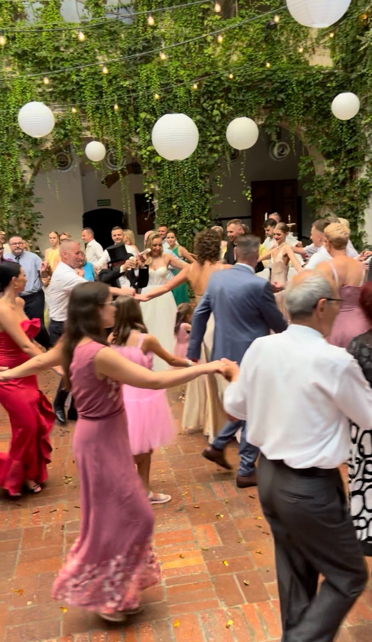 Radosny korowód weselny w plenerze: tańczący goście w kolorowych strojach, panna młoda w bieli, dekoracje z lampionów i zieleni tworzą unikalny klimat zabawy.