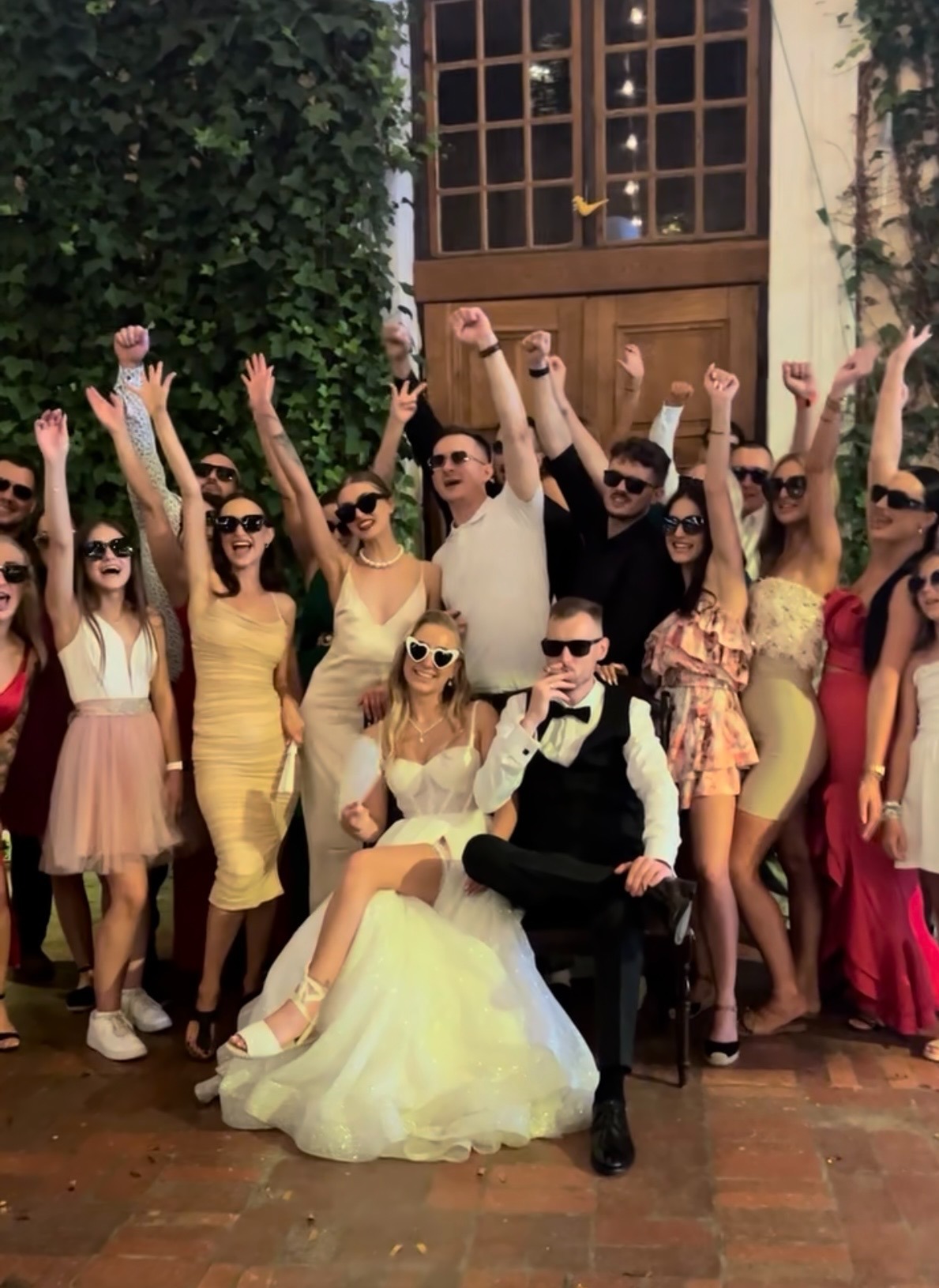 Radosna grupa gości weselnych z parą młodą w centrum, wszyscy w okularach przeciwsłonecznych, pozują z uniesionymi rękami na tle budynku porośniętego bluszczem.