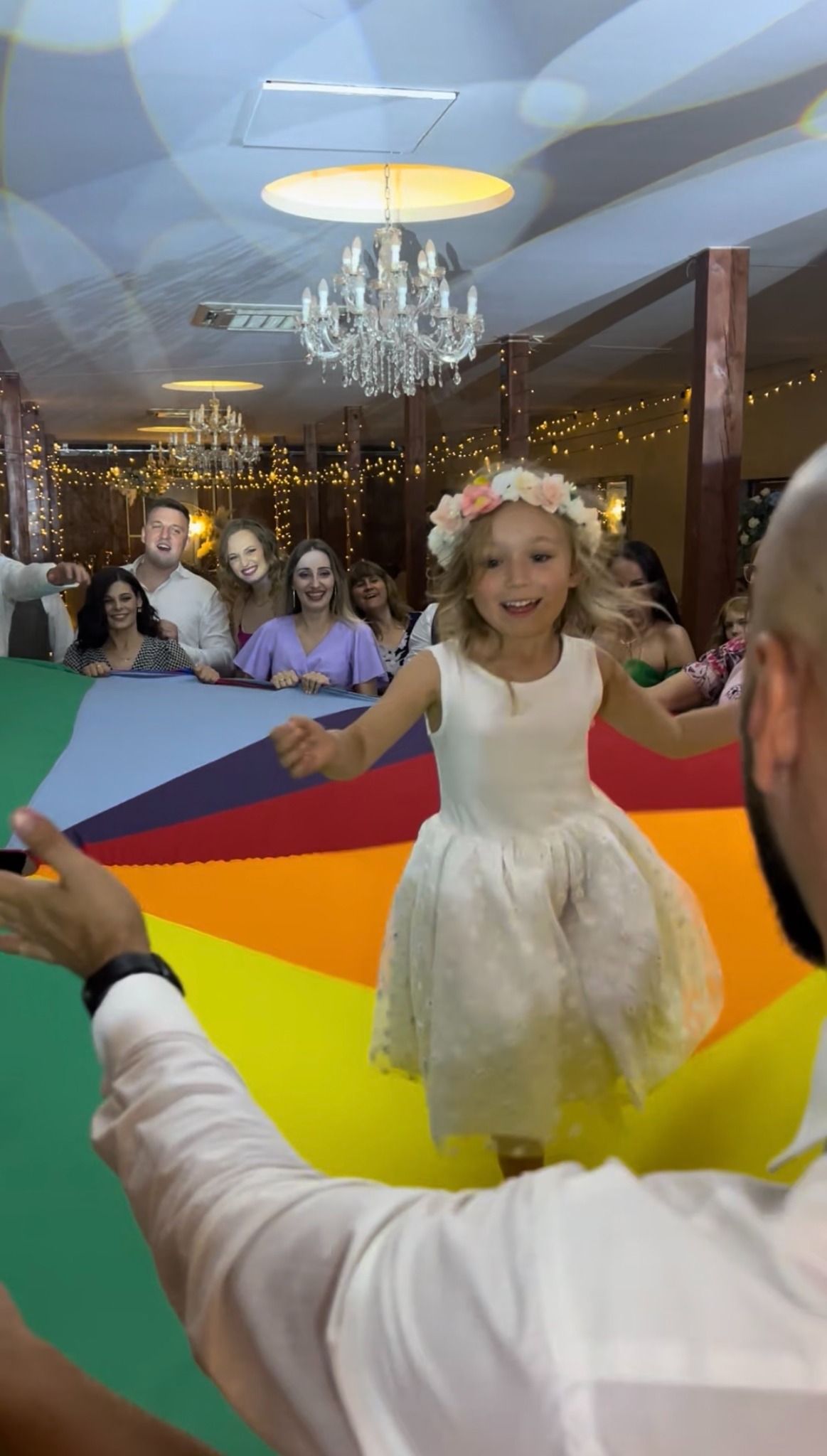 Dziewczynka w białej sukience tańczy na kolorowej chuście podczas imprezy w Międzyrzeczu. W tle uśmiechnięci goście i dekoracje.
