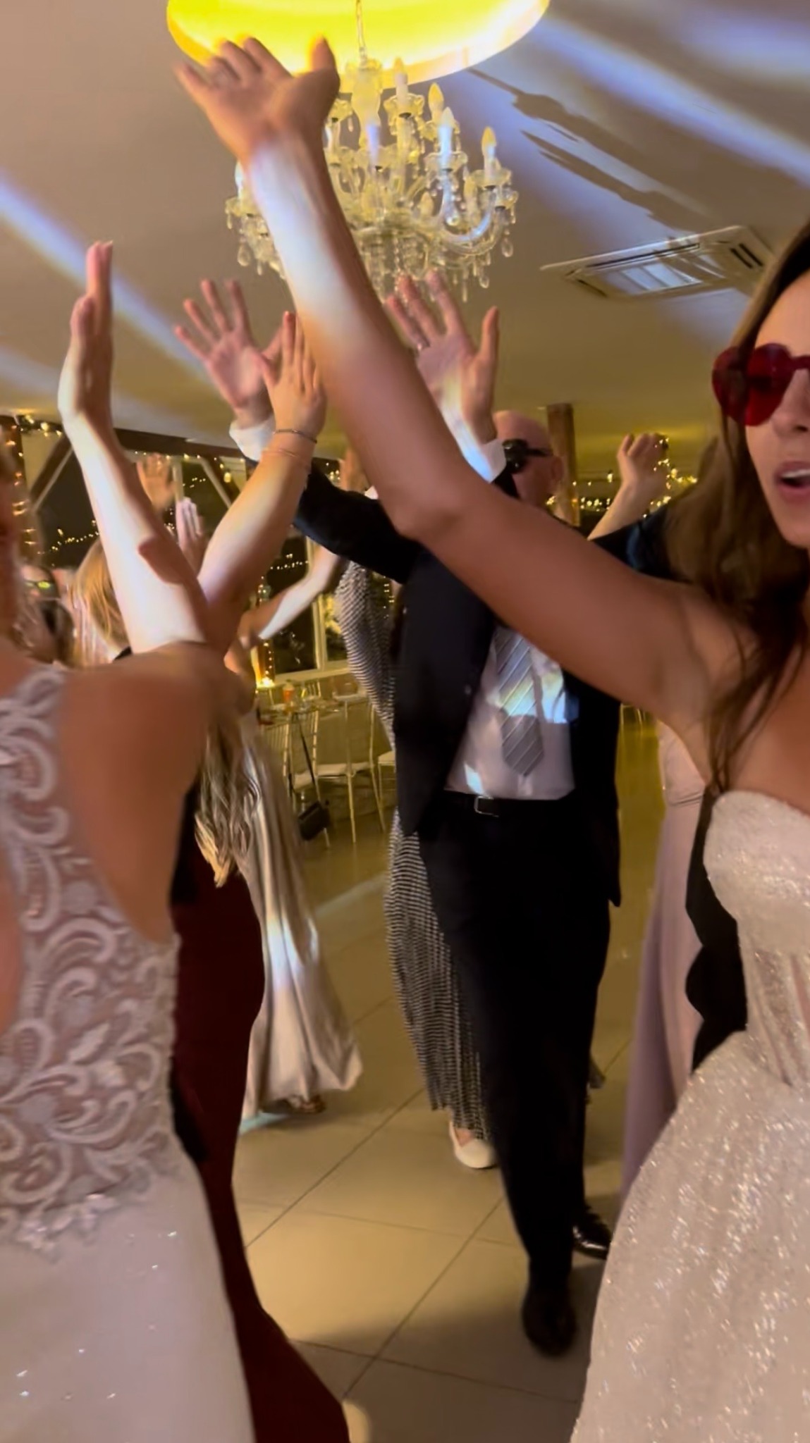 Roztańczona grupa gości weselnych w eleganckich strojach, wznoszą ręce w górę podczas zabawy na parkiecie, oświetlonym przez dekoracyjny żyrandol.