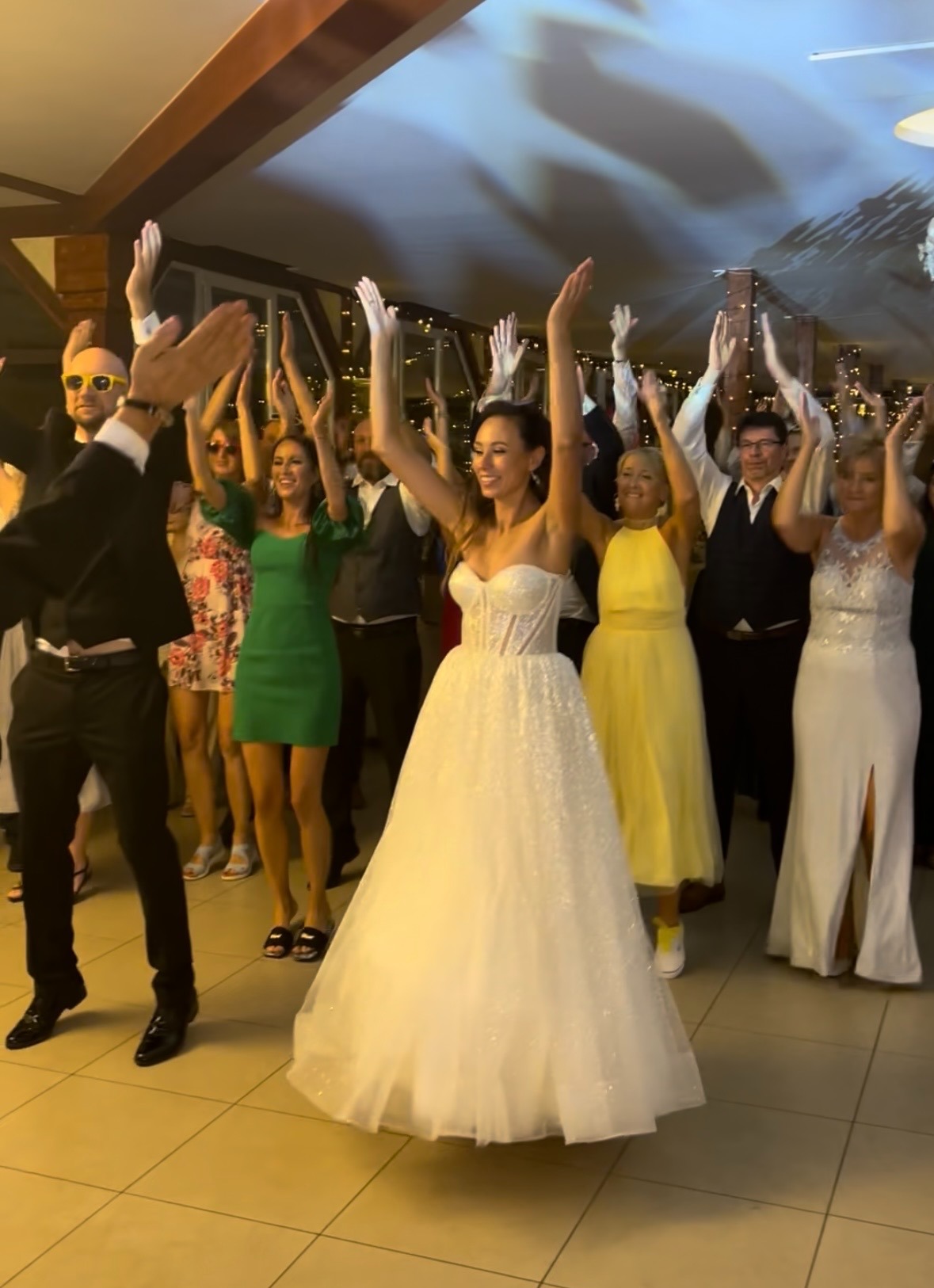 Roztańczona grupa gości weselnych z podniesionymi rękami, w centrum panna młoda w białej sukni. Ujęcie z parkietu, ciepłe oświetlenie.