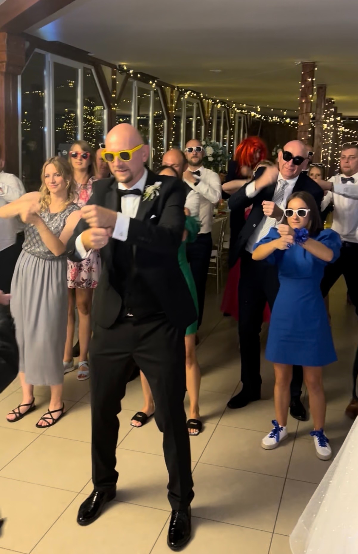 Rozbawieni goście weselni w okularach tańczą w rytm muzyki. W tle dekoracje świetlne i okna. Ujęcie z perspektywy parkietu.