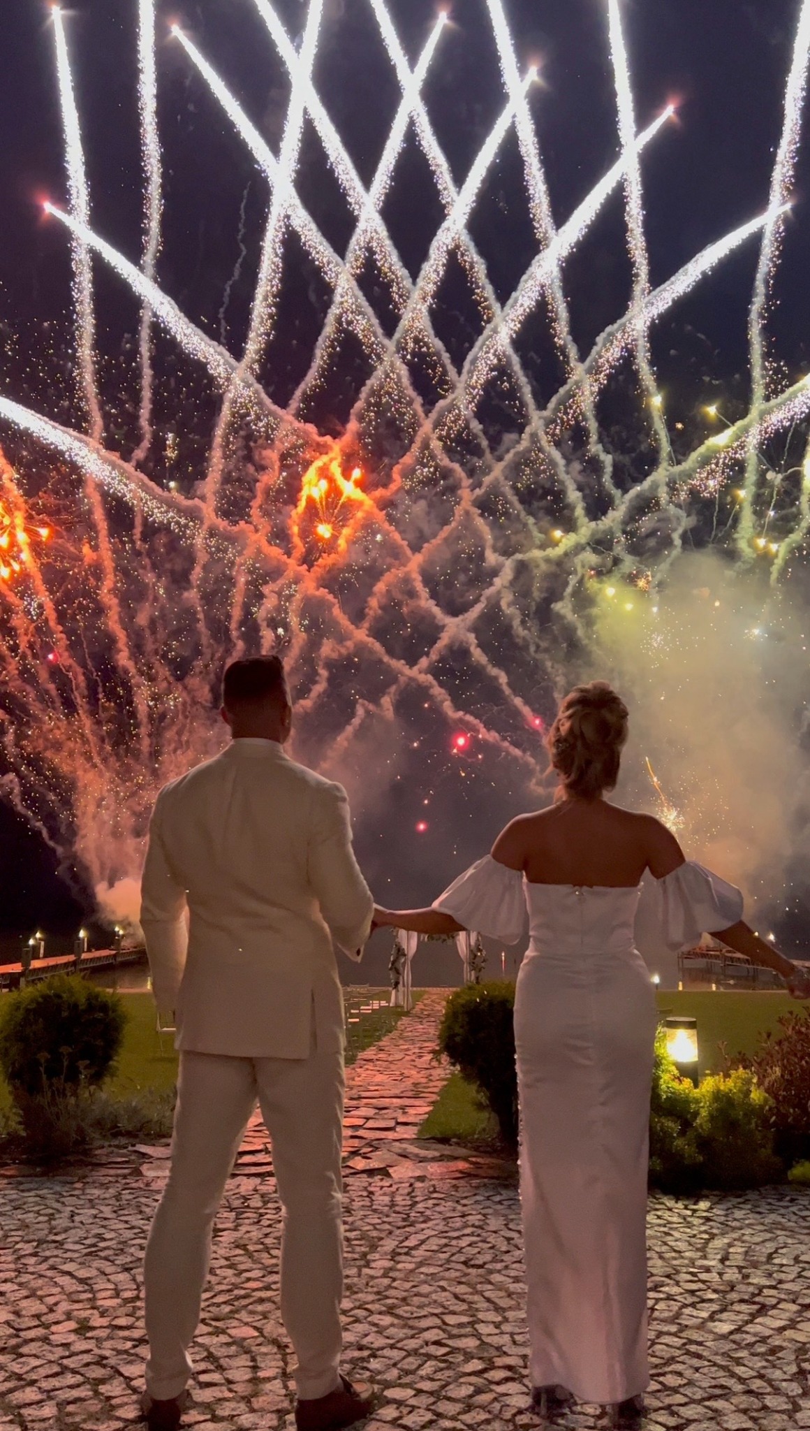 Para młoda w białych strojach podziwia pokaz sztucznych ogni nad kamienną ścieżką, wieczorową porą. Widok z tyłu, oświetlenie sceniczne.