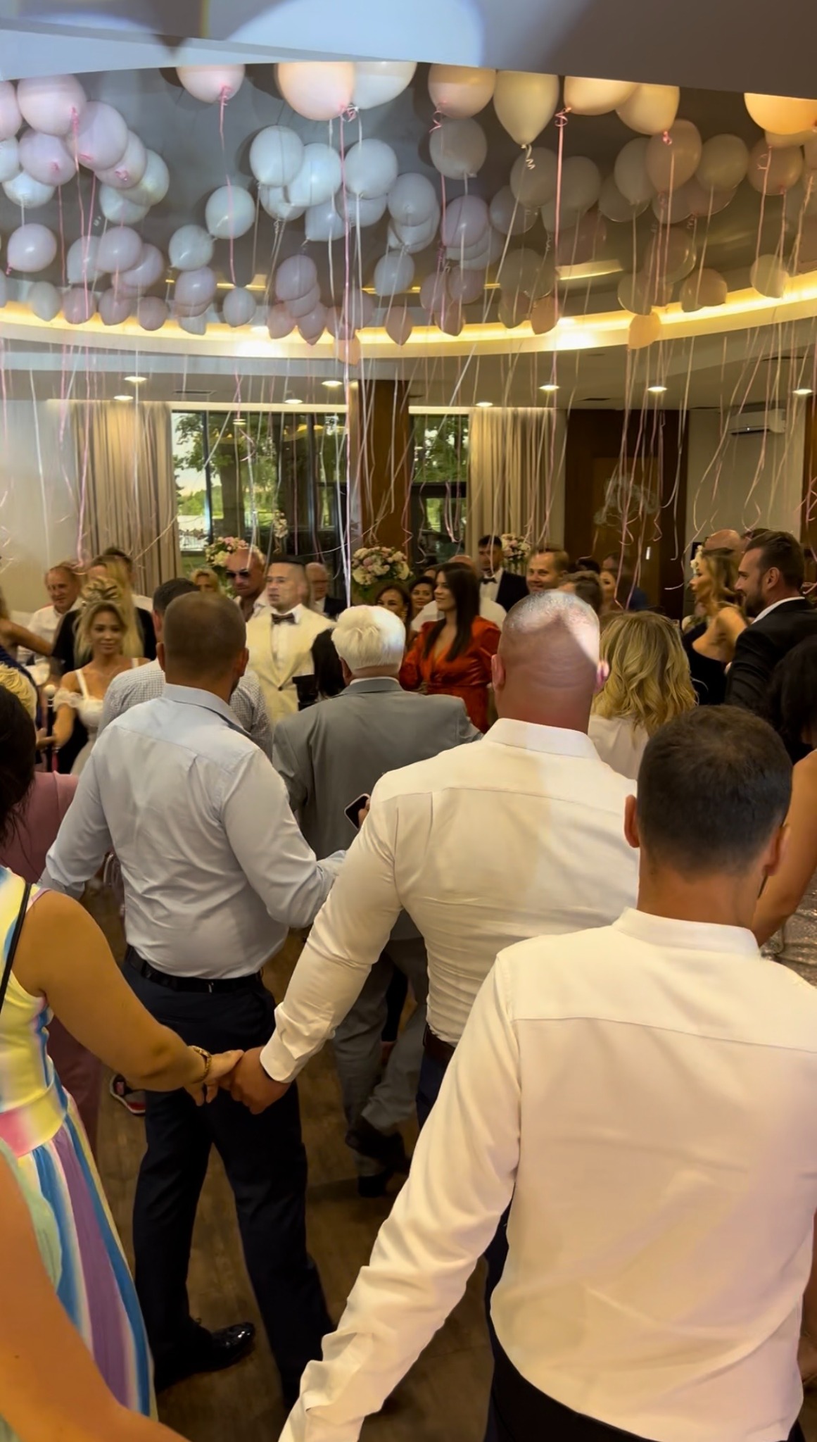Goście weselni trzymają się za ręce, tworząc krąg na parkiecie, nad nimi dekoracja z białych i różowych balonów. W tle widoczni inni goście i dekoracje kwiatowe.
