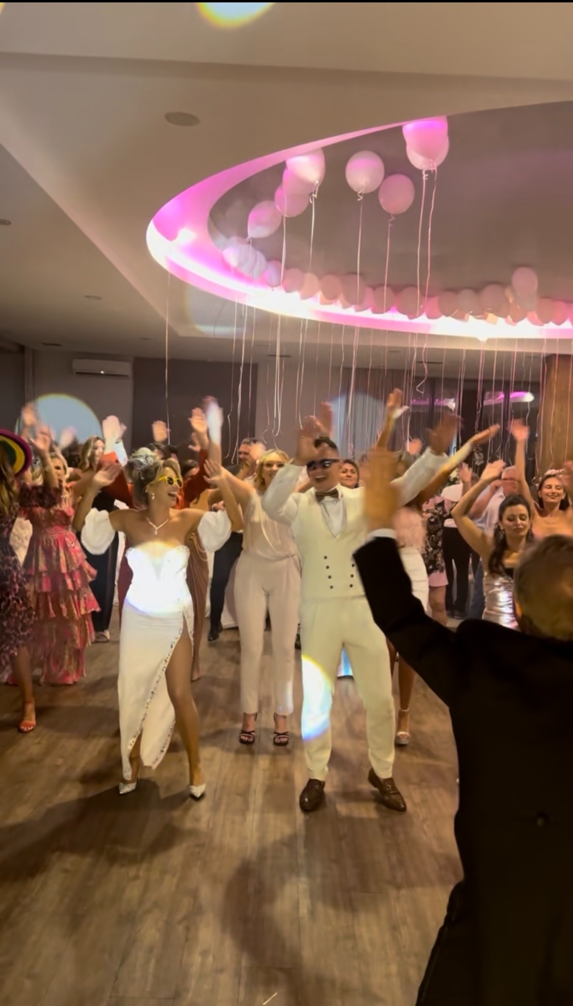 Rozbawieni goście weselni tańczą na parkiecie pod dekoracją z białych balonów i różowym oświetleniem sufitowym. W centrum para młoda w białych strojach.