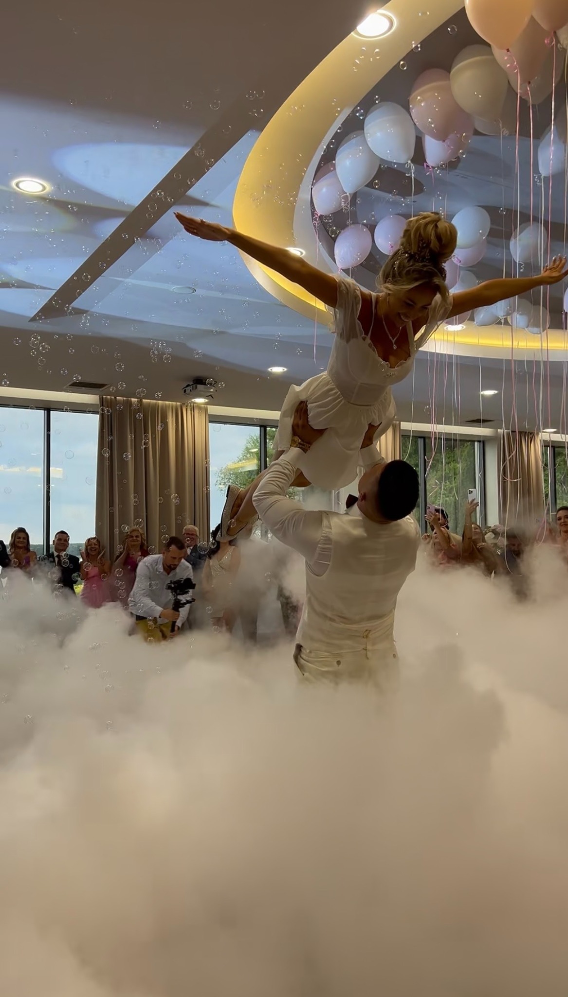 Para w białych strojach tańczy w dymie na tle gości i dekoracji balonowych. Mężczyzna podnosi kobietę w górę. Ujęcie z imprezy.