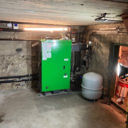 GB-INSTAL TERMO Grzegorz Bystrzyk - Zielony piec grzewczy w piwnicy z rurami, zbiornikiem wyrównawczym i lampą oświetleniową. Surowy betonowy strop i ściany. Instalacja połączona z siecią.