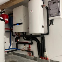 GB-INSTAL TERMO Grzegorz Bystrzyk - Złożona instalacja grzewcza z białym zasobnikiem, kotłem i rozdzielnicą elektryczną na ścianie. Różnokolorowe rury i czarne izolacje tworzą sieć połączeń.