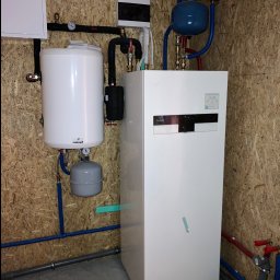 GB-INSTAL TERMO Grzegorz Bystrzyk - Biała pompa ciepła Panasonic Aquarea Smart Cloud z zbiornikiem Galmet, zainstalowana na ścianie z płyty OSB, z widocznymi rurami instalacji hydraulicznej.