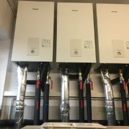 Trzy białe jednostki Panasonic Aquarea Smart Cloud zamontowane na ścianie, z izolowanymi rurami i zaworami, widoczne strzałki na rurach wskazujące kierunek przepływu.