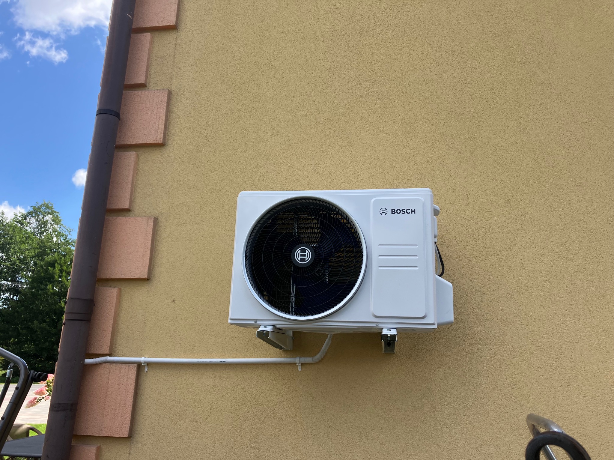 Biała jednostka zewnętrzna klimatyzacji Bosch zamontowana na beżowej ścianie budynku. Widoczny fragment rynny i niebieskie niebo z chmurami w tle.