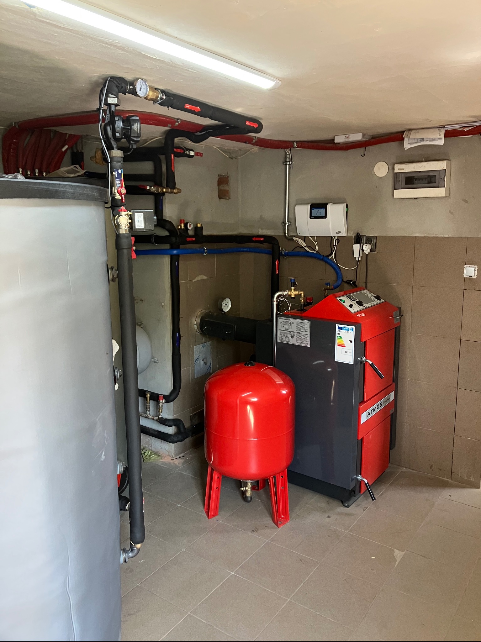 Zainstalowany, kompletny system grzewczy z kotłem Atmos, zbiornikiem akumulacyjnym i czerwonym naczyniem wzbiorczym w kotłowni z wykafelkowanymi ścianami.