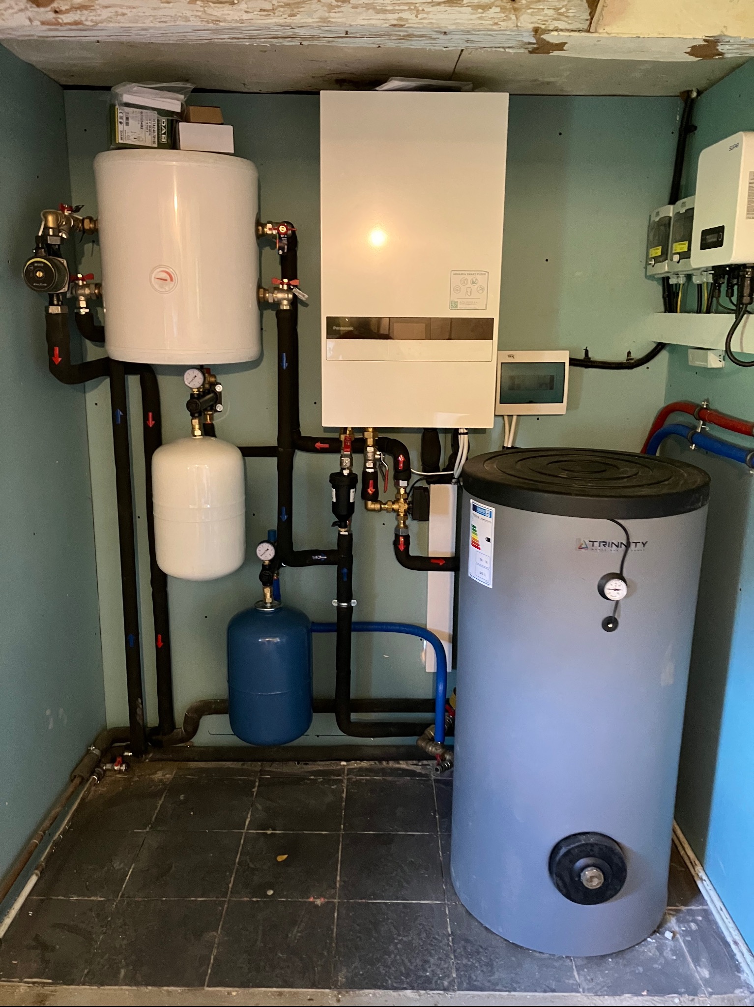 Zestaw instalacji hydraulicznej z zbiornikami, rurami i zaworami w pomieszczeniu z niebieskimi ścianami i czarną podłogą. Widoczny bojler Trinity i inne komponenty.