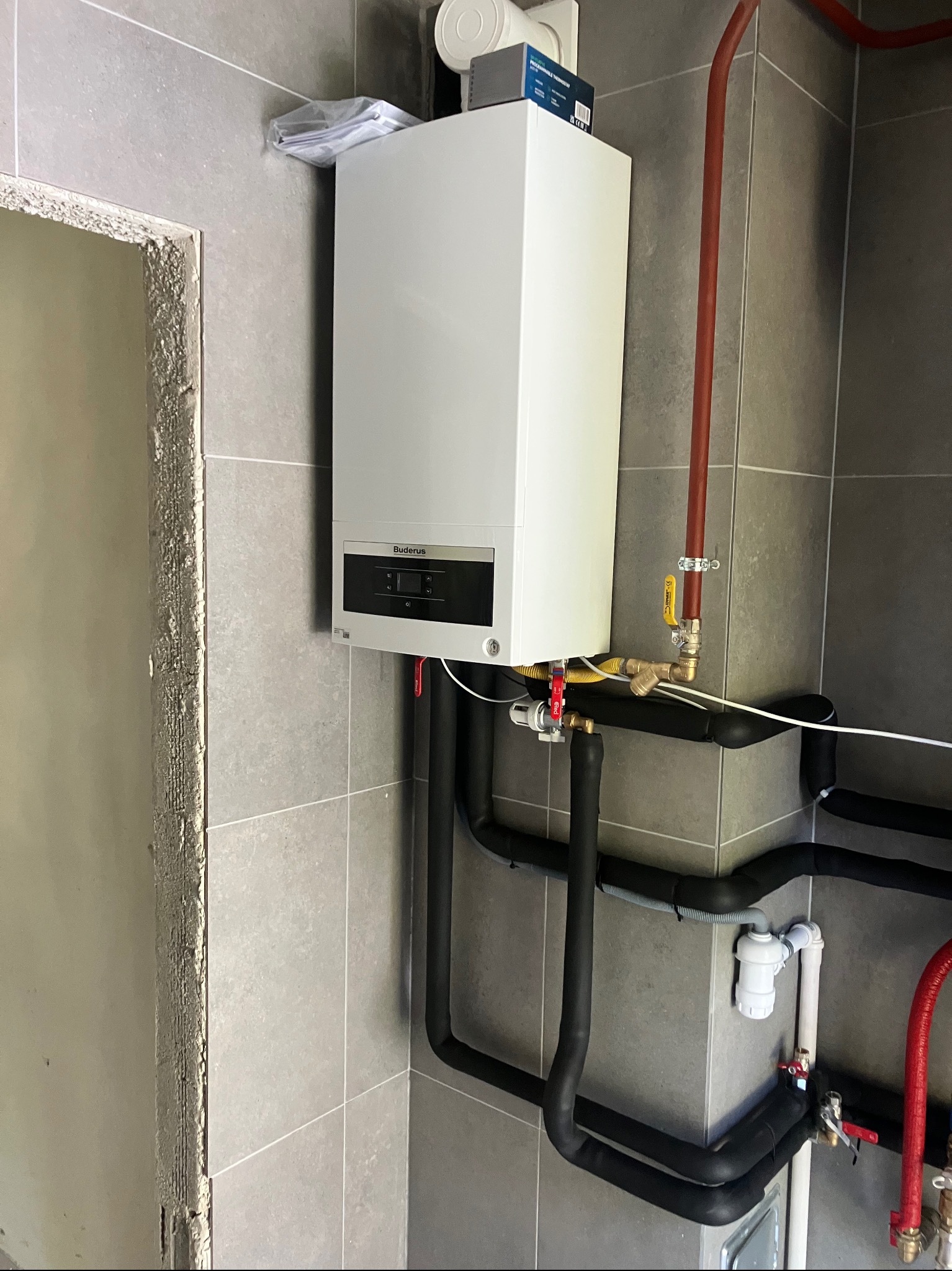 Biały piec gazowy Buderus zamontowany na szarej, kafelkowanej ścianie w Kolbuszowej. Widoczne rury instalacji grzewczej i fragment surowej ściany obok.