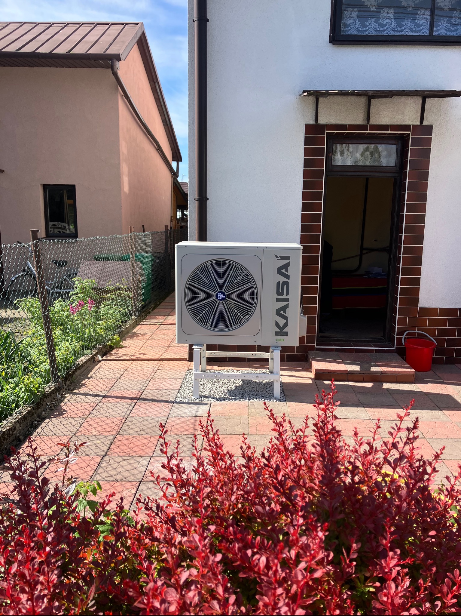 Zamontowana pompa ciepła Kaisai na zewnątrz budynku, widoczna z poziomu gruntu, z czerwonymi krzewami na pierwszym planie i budynkami w tle.