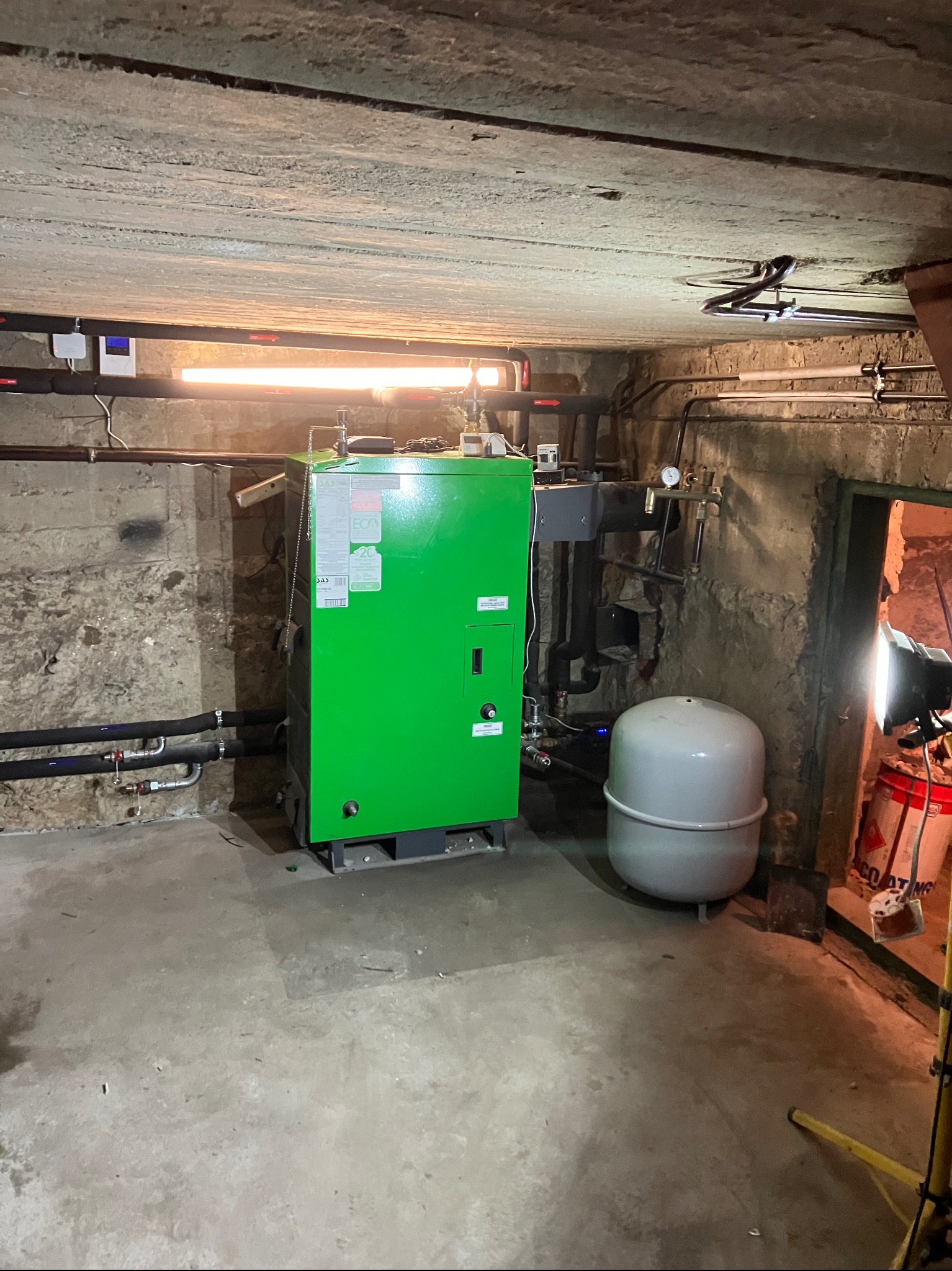 Zielony piec grzewczy w piwnicy z rurami, zbiornikiem wyrównawczym i lampą oświetleniową. Surowy betonowy strop i ściany. Instalacja połączona z siecią.