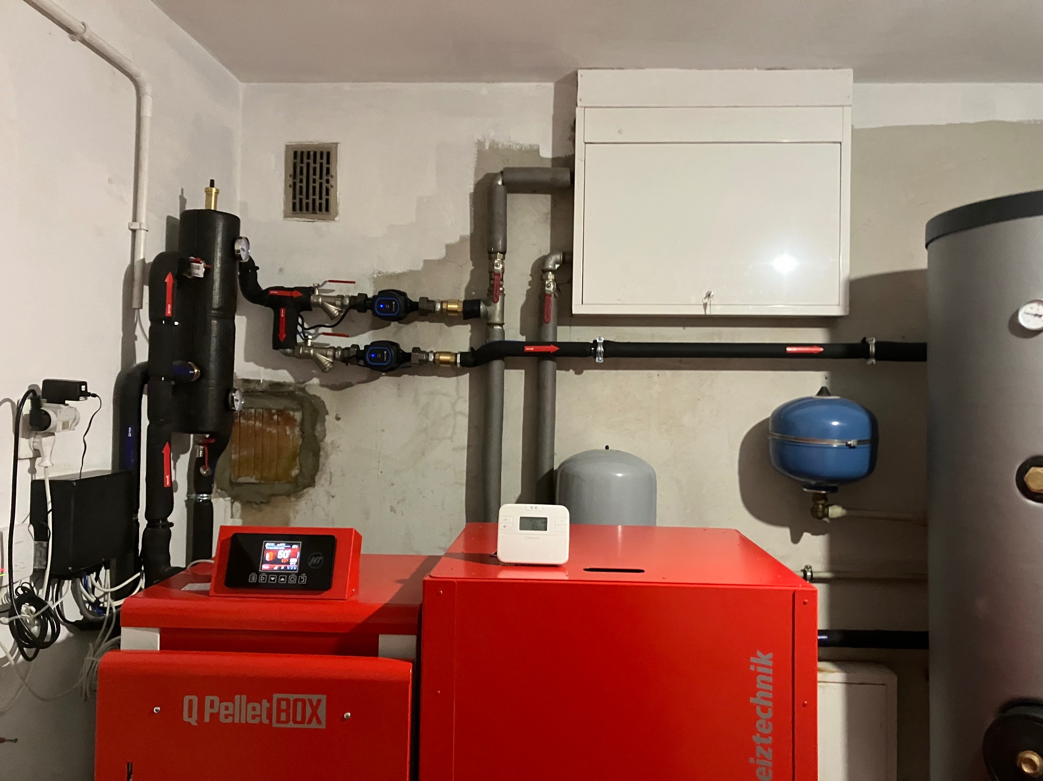 Czerwony piec na pellet Q Pellet BOX w kotłowni, z widocznymi rurami, zbiornikiem i sterownikiem. Elementy instalacji grzewczej w pomieszczeniu.