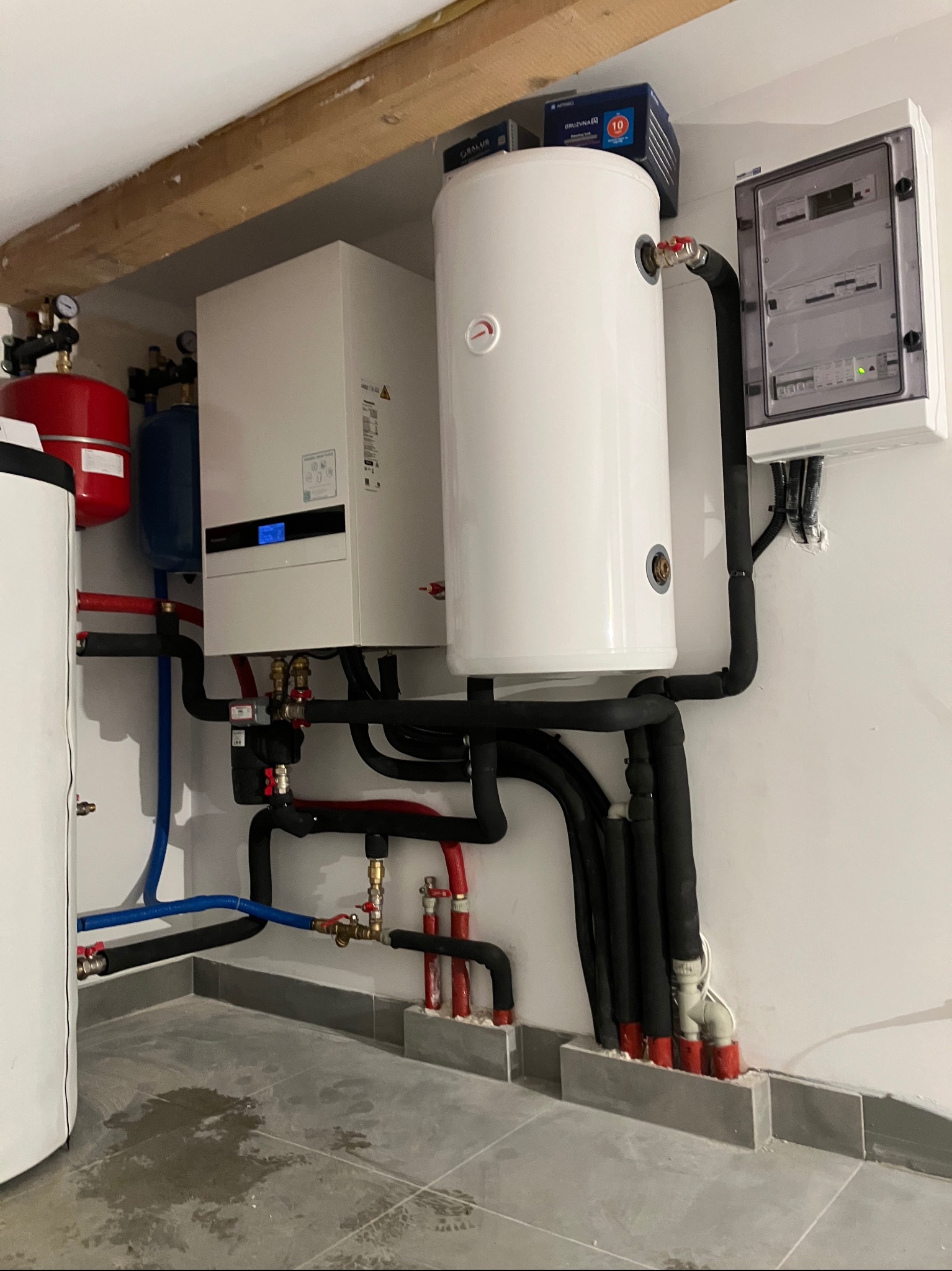 Złożona instalacja grzewcza z białym zasobnikiem, kotłem i rozdzielnicą elektryczną na ścianie. Różnokolorowe rury i czarne izolacje tworzą sieć połączeń.