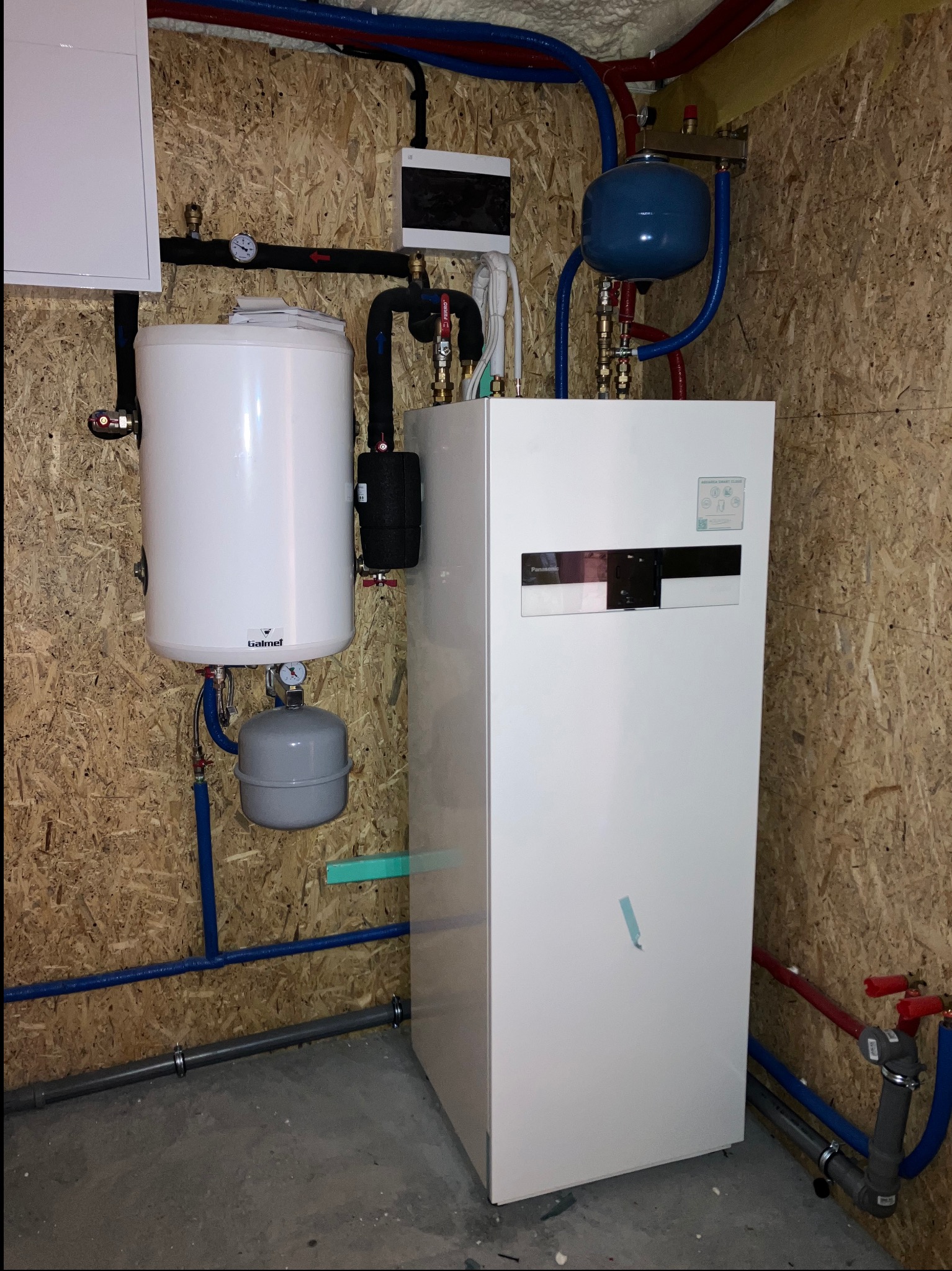 Biała pompa ciepła Panasonic Aquarea Smart Cloud z zbiornikiem Galmet, zainstalowana na ścianie z płyty OSB, z widocznymi rurami instalacji hydraulicznej.