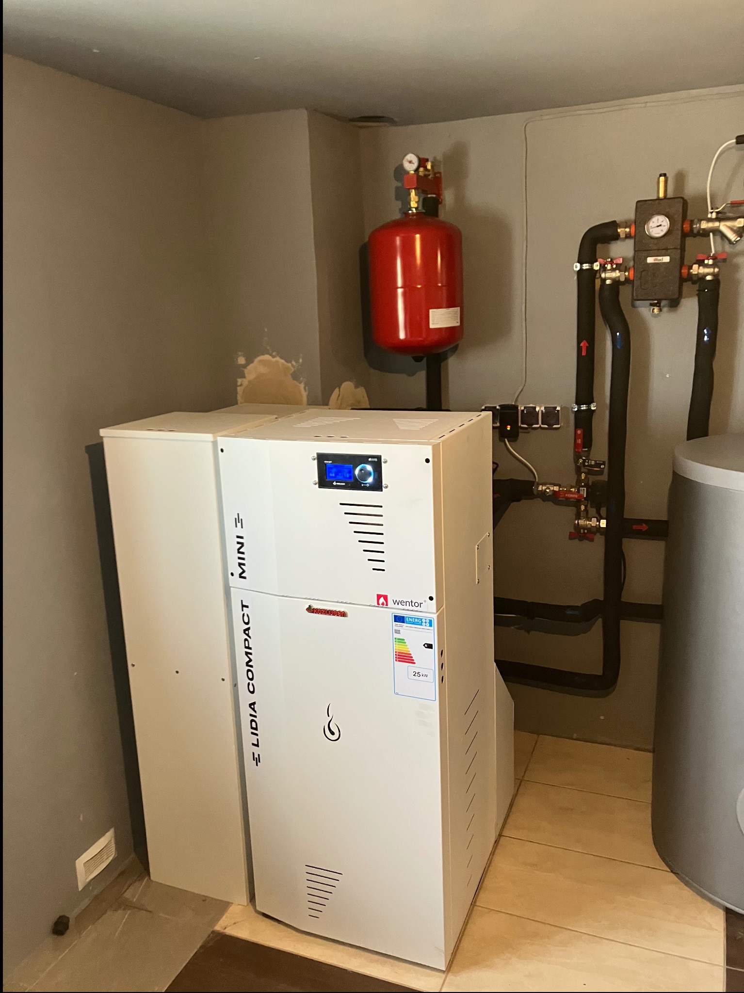 Kompaktowy piec grzewczy Lidia Compact Mini z podłączonymi instalacjami, w tym czerwonym zbiornikiem wyrównawczym i czarnymi rurami, zamontowany w szarej kotłowni.