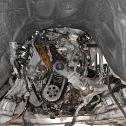 Widok od dołu na silnik samochodowy w trakcie naprawy, z widocznym łańcuchem rozrządu, elementami układu smarowania i przewodami, w warsztatowym otoczeniu.