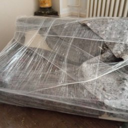 AR-TRANS Agata Rudnicka - Sofa owinięta folią stretch i zabezpieczona kocem transportowym, przygotowana do przeniesienia, widoczna na tle drewnianej podłogi i grzejnika.