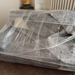 Sofa owinięta folią stretch i zabezpieczona kocem transportowym, przygotowana do przeniesienia, widoczna na tle drewnianej podłogi i grzejnika.