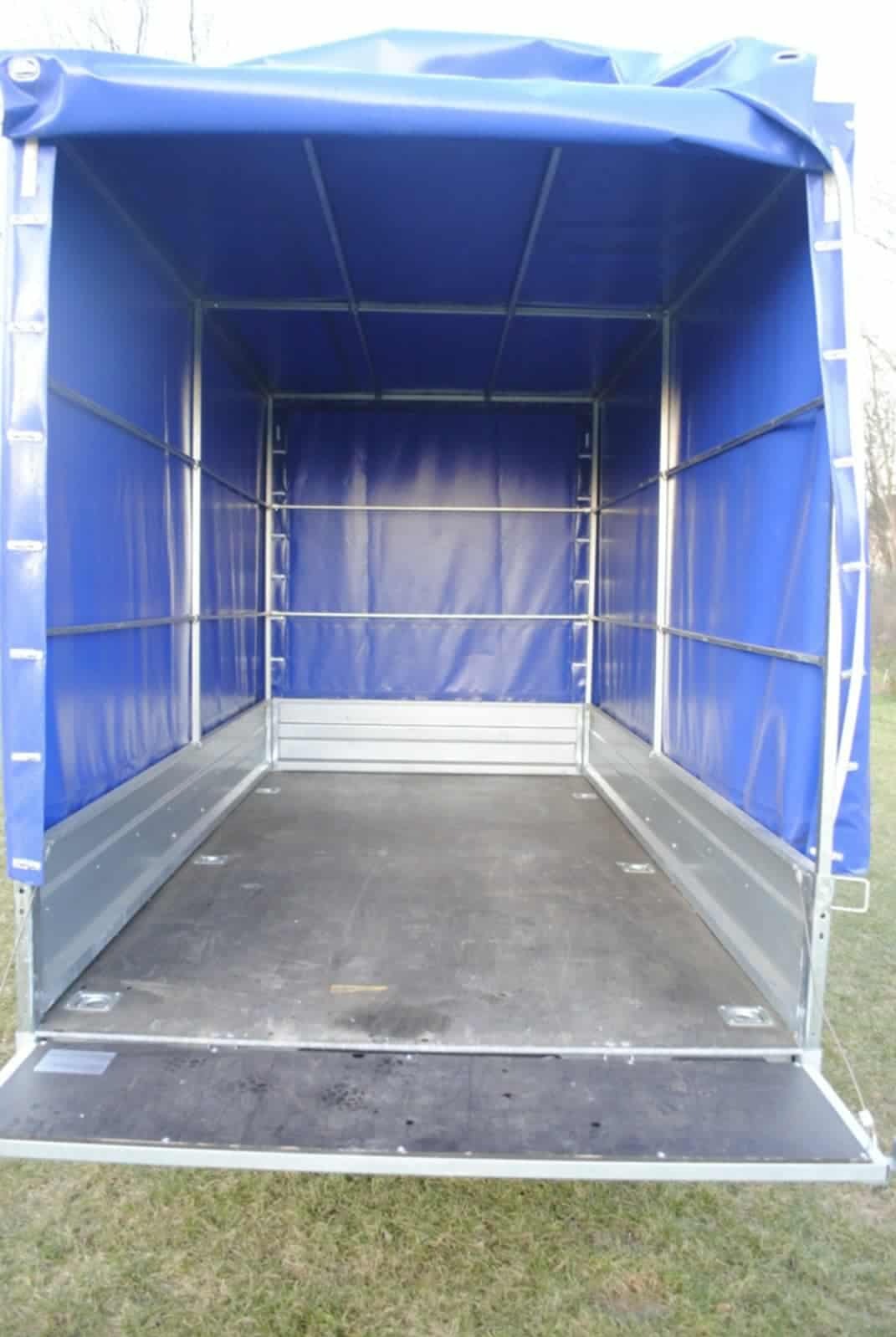 Plandeka z otwartą klapą załadunkową. Wnętrze puste, gotowe do załadunku. Widoczne mocowania i metalowa konstrukcja. Trawa w tle.