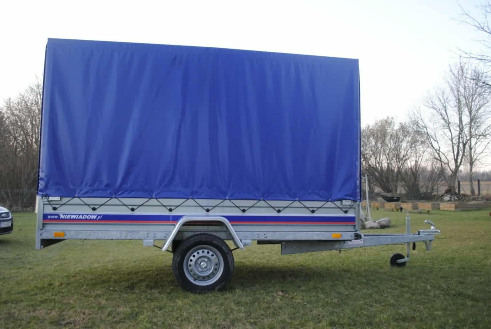 Przyczepa Niewiadów z plandeką w kolorze niebieskim, zaparkowana na trawie. Widoczny fragment samochodu osobowego w tle. Przyczepa gotowa do transportu.