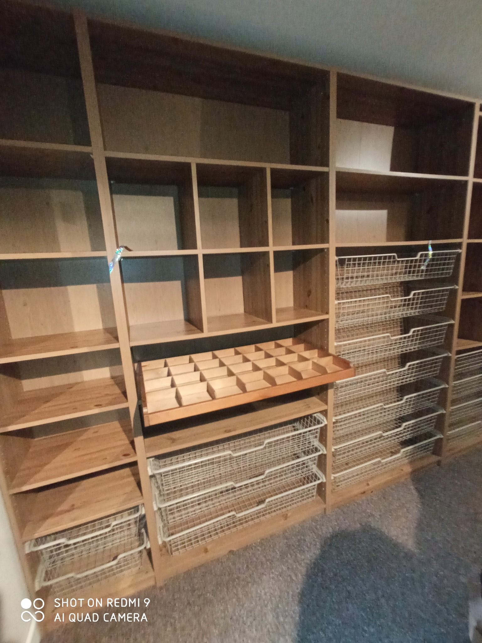 Drewniany regał z półkami i wysuwanymi koszami z metalowej siatki, widoczny fragment podłogi pokrytej wykładziną.