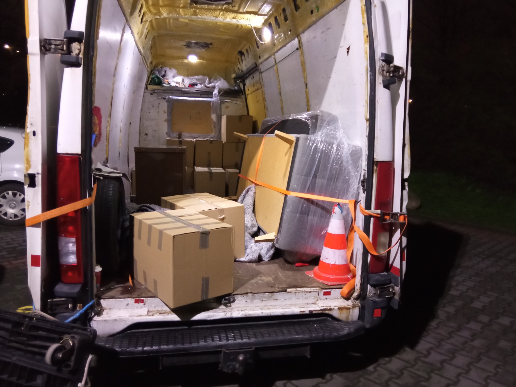 Wnętrze częściowo załadowanego busa z kartonami, meblami owiniętymi folią i pomarańczowym pachołkiem, zabezpieczone pomarańczowymi pasami transportowymi, widoczne oświetlenie wewnętrzne.