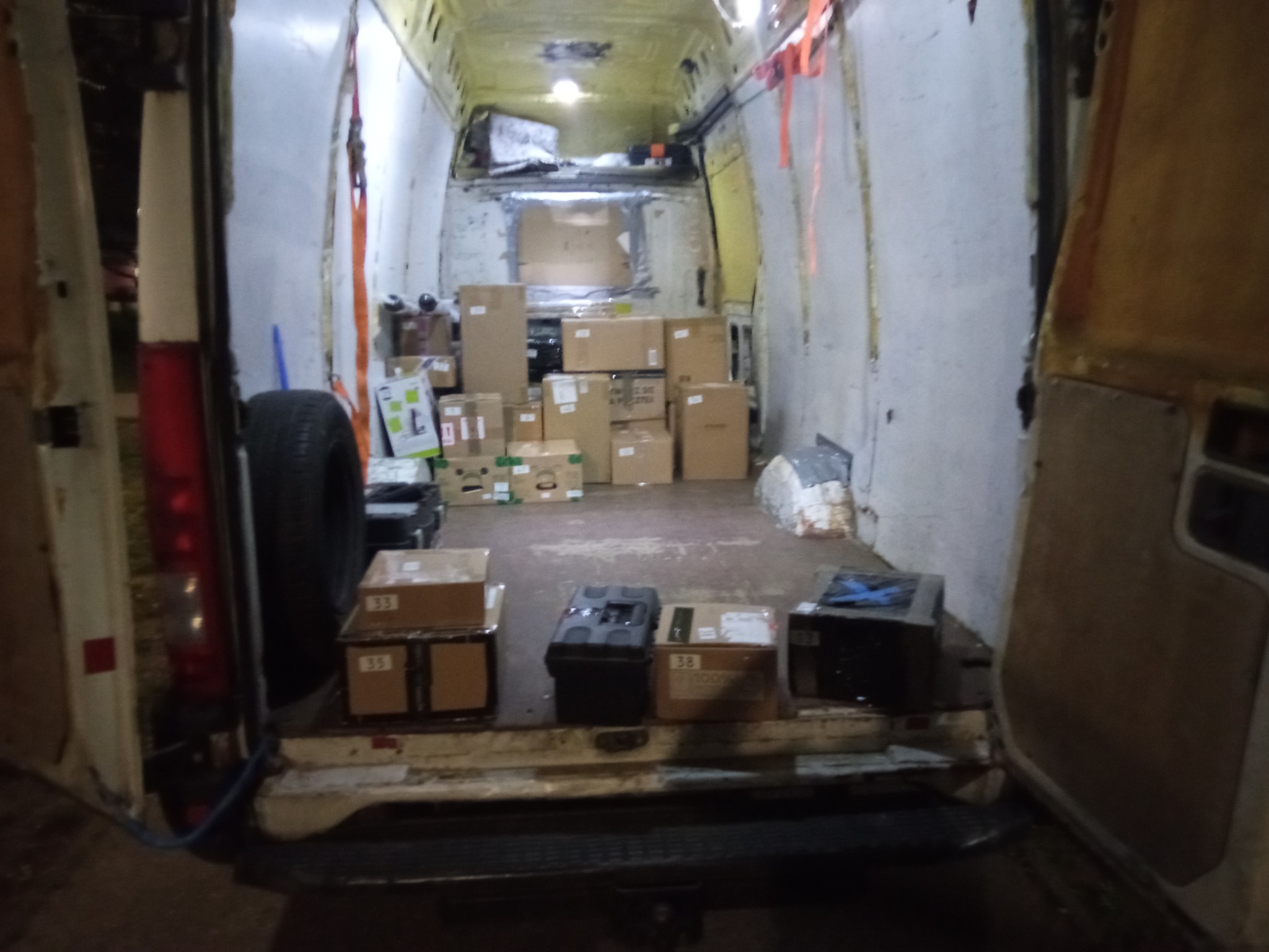 Wnętrze białego, częściowo załadowanego busa z otwartymi drzwiami, widoczne kartony, opona i zabezpieczenia ładunku.