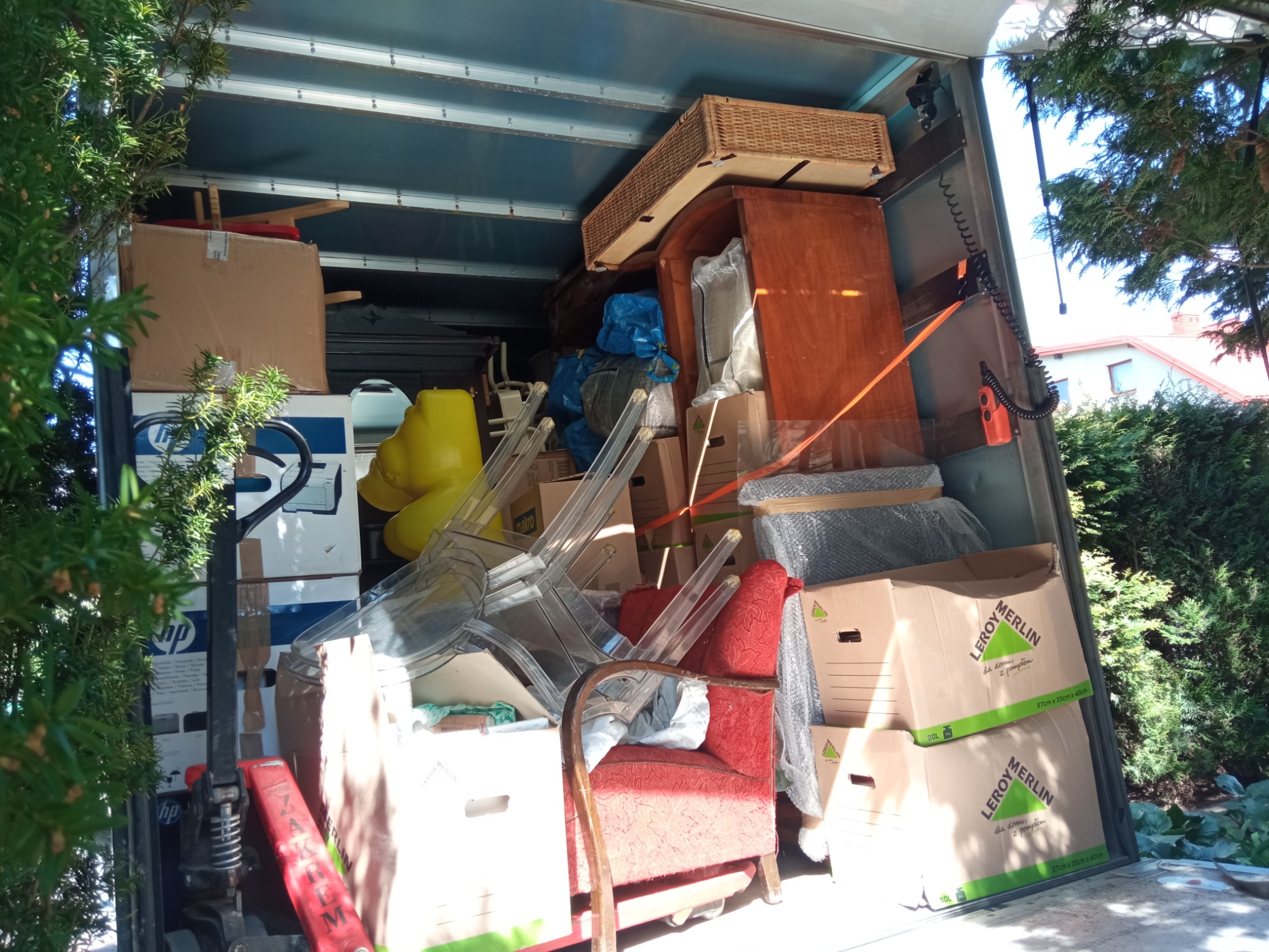 Wnętrze załadowanego busa przeprowadzkowego z meblami (czerwony fotel, przezroczyste krzesła), kartonami (w tym Leroy Merlin) i sprzętem, widok z zewnątrz przez otwarte drzwi, częściowo zasłonięte...