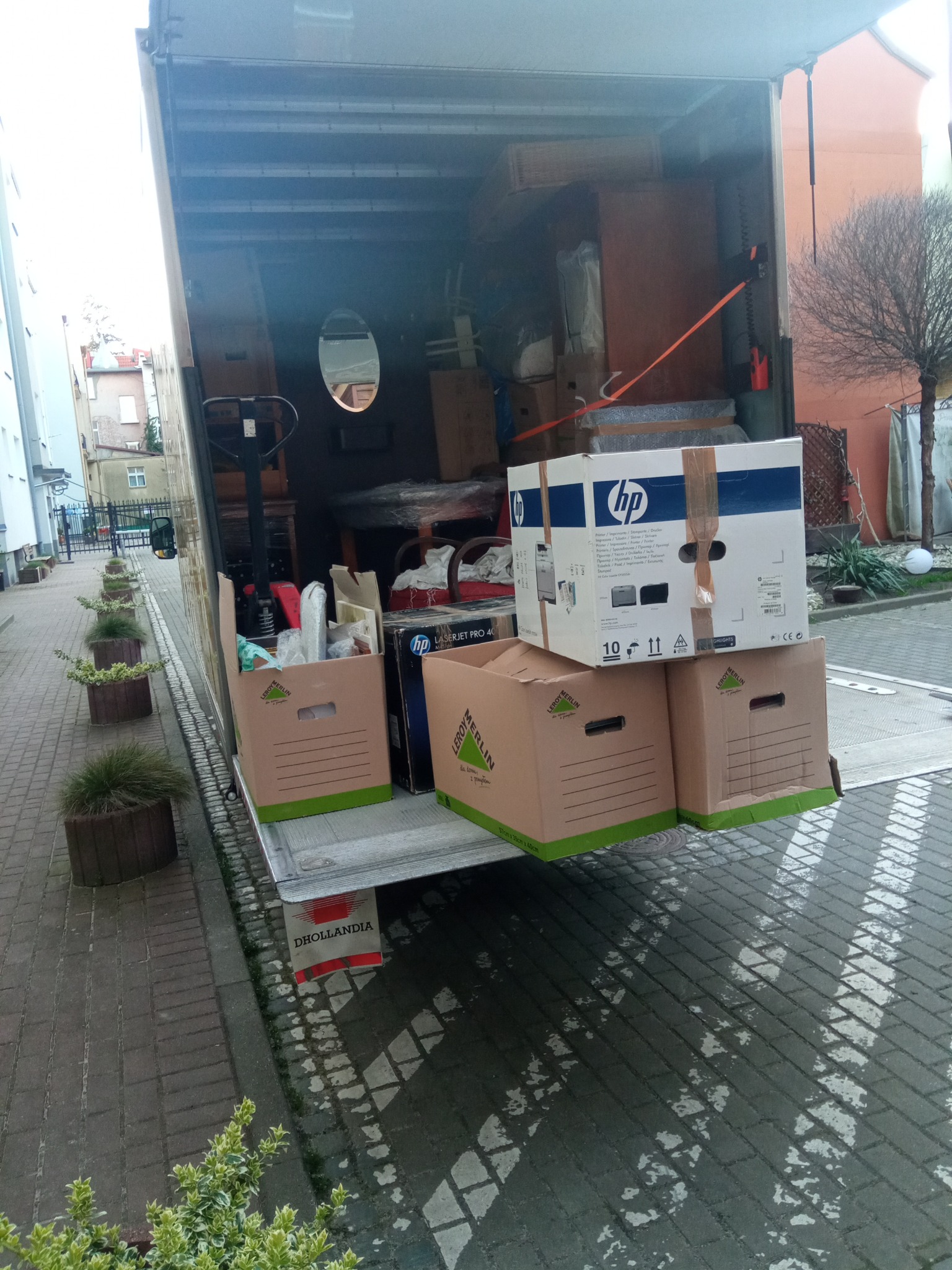 Otwarta paka samochodu dostawczego załadowana meblami i kartonami, w tym pudełkami Leroy Merlin i HP, widoczny wózek transportowy wewnątrz, tło miejskie z budynkami i roślinnością, mokra kostka...