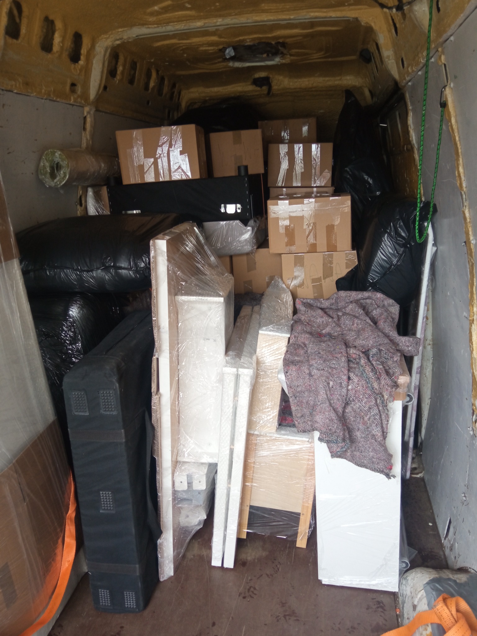 Wnętrze furgonetki załadowane kartonami, meblami owiniętymi folią, czarnymi workami i kocem zabezpieczającym ładunek podczas transportu.