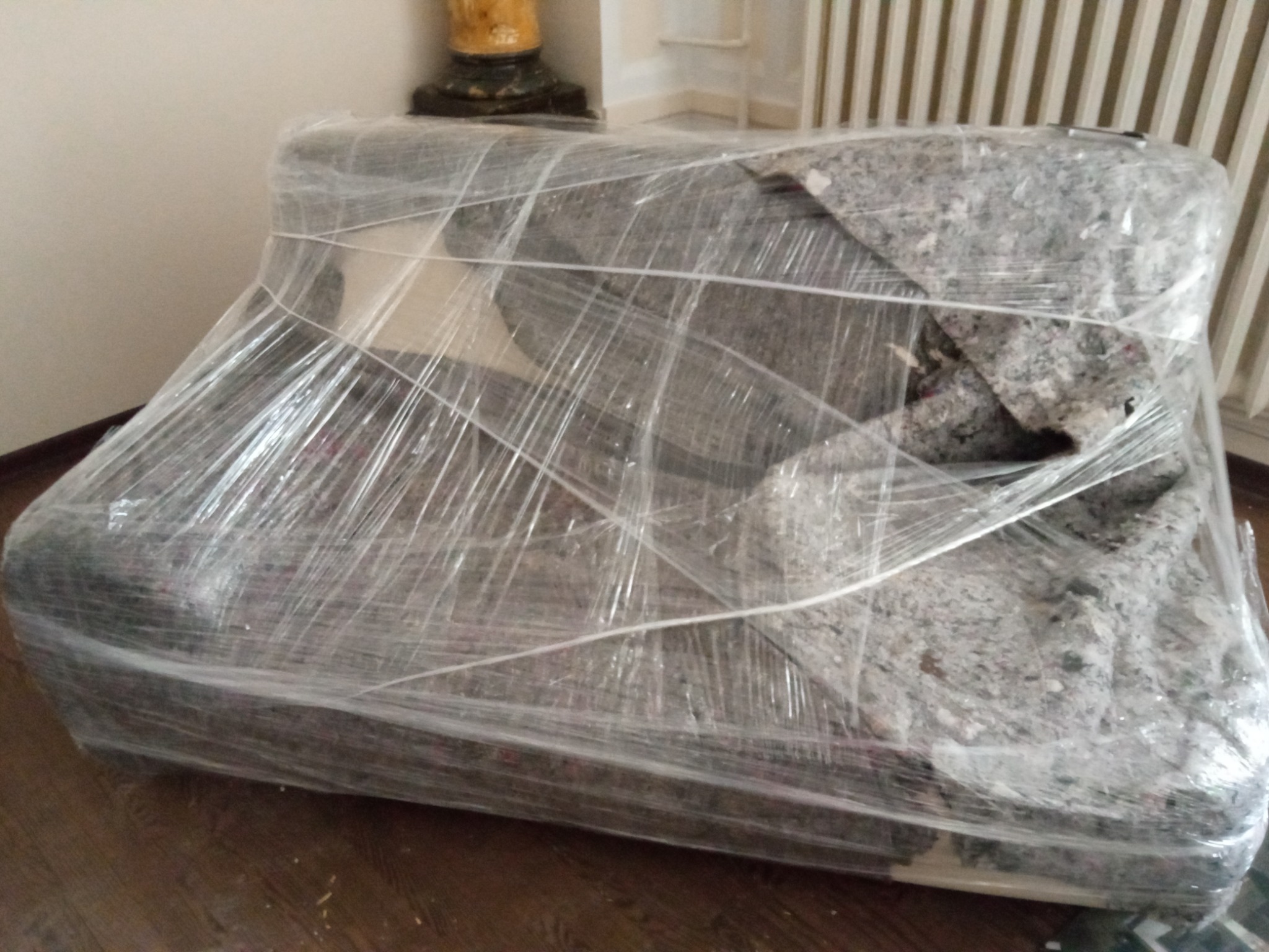 Sofa owinięta folią stretch i zabezpieczona kocem transportowym, przygotowana do przeniesienia, widoczna na tle drewnianej podłogi i grzejnika.