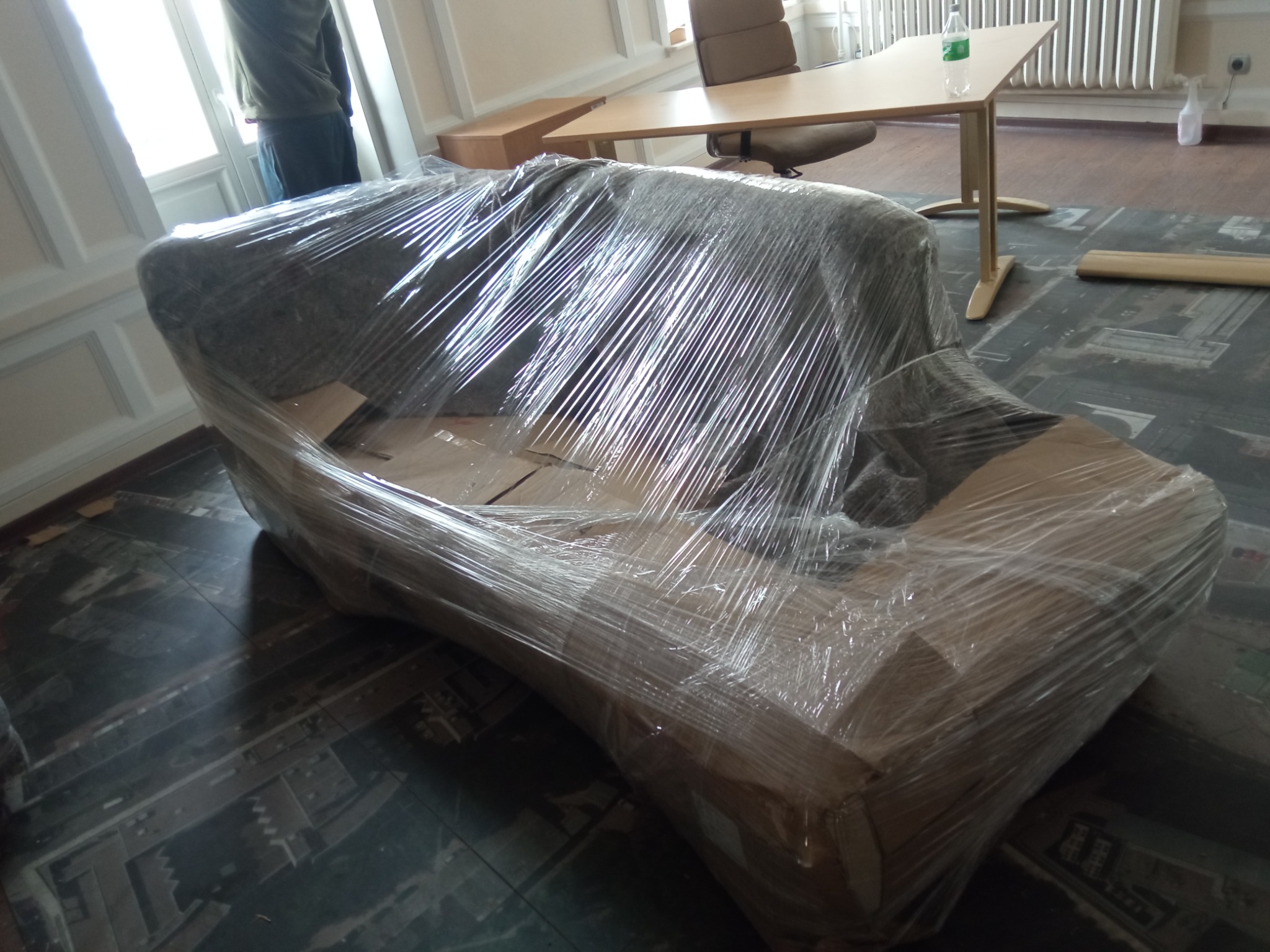 Sofa owinięta folią stretch, przygotowana do transportu w pomieszczeniu z biurkiem i krzesłem w tle.